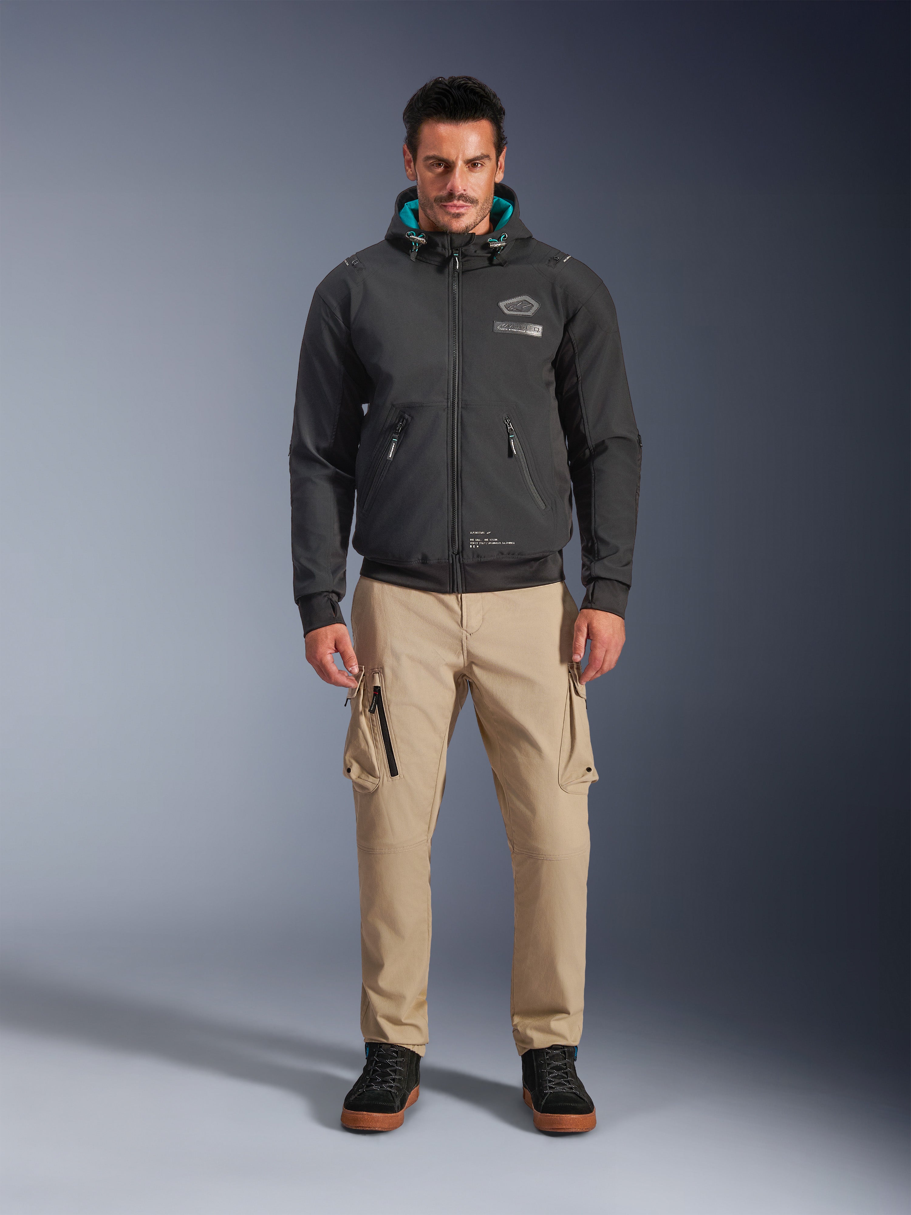 Alpinestars Moblast Waterproof Jacket, Black urban motorcycle hoodie with teal interior hood lining, worn with tan cargo pants and black high-top boots, contemporary all-season riding apparel