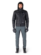 Alpinestars Mohobbs Wasserdichte Jacke in Schwarz, männliches Model in gesteppter wattierter Bomberjacke mit Kapuze, graue Motorradhose, schwarze Handschuhe und schwarze Stiefel, urbane zeitgenössische Motorradausrüstung.