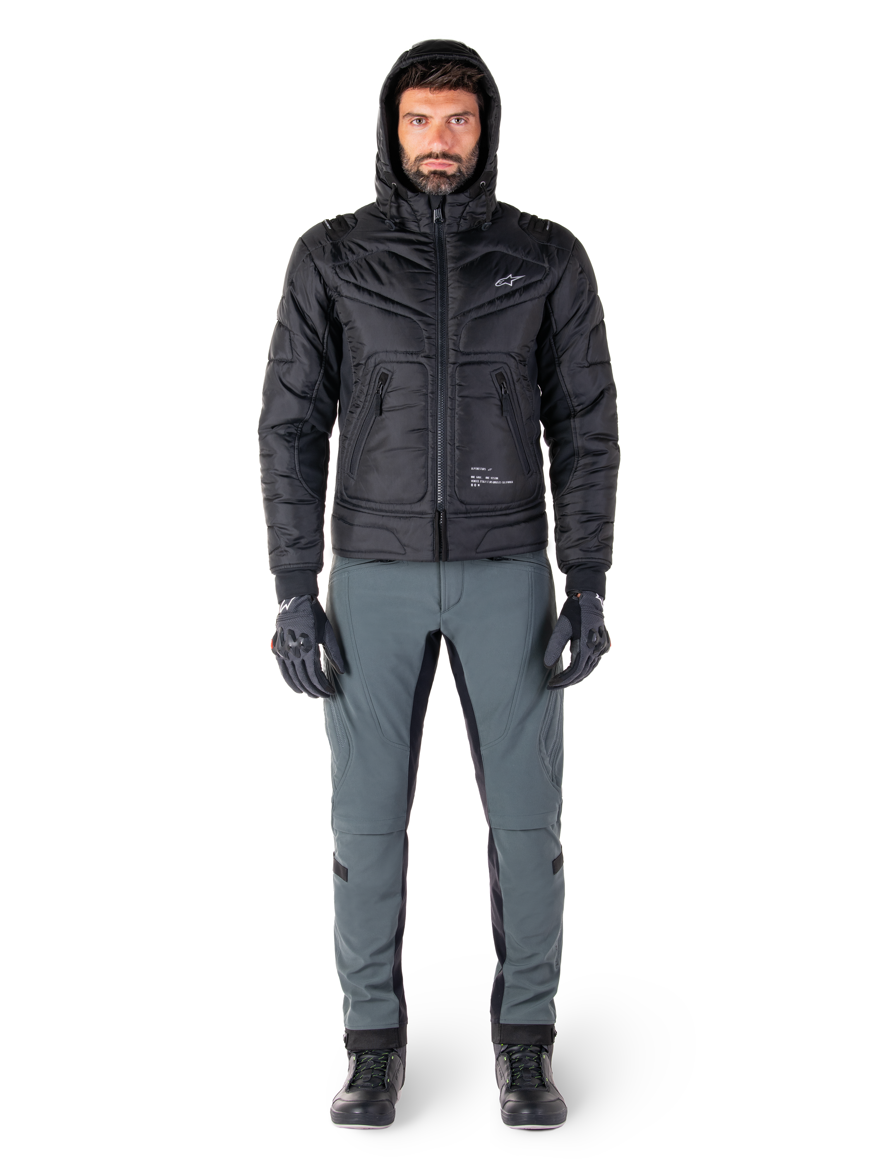 Alpinestars Mohobbs Wasserdichte Jacke in Schwarz, männliches Model in gesteppter wattierter Bomberjacke mit Kapuze, graue Motorradhose, schwarze Handschuhe und schwarze Stiefel, urbane zeitgenössische Motorradausrüstung.