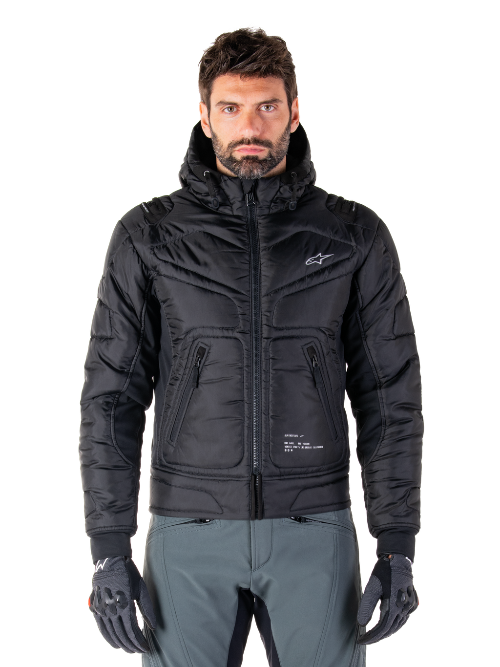 Alpinestars Mohobbs Waterproof Jacket, schwarze gepolsterte Bomber-Motorradjacke, gestepptes Polygewebe mit Frontreißverschluss und Reißverschlusstaschen, kleines weißes Logo auf der Brust, Modell trägt graue Hose und Handschuhe, urbanes Streetwear-Design für Stadtfahrten
