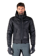 Alpinestars Mohobbs Waterproof Jacket, schwarze gepolsterte Bomber-Motorradjacke, gestepptes Polygewebe mit Frontreißverschluss und Reißverschlusstaschen, kleines weißes Logo auf der Brust, Modell trägt graue Hose und Handschuhe, urbanes Streetwear-Design für Stadtfahrten
