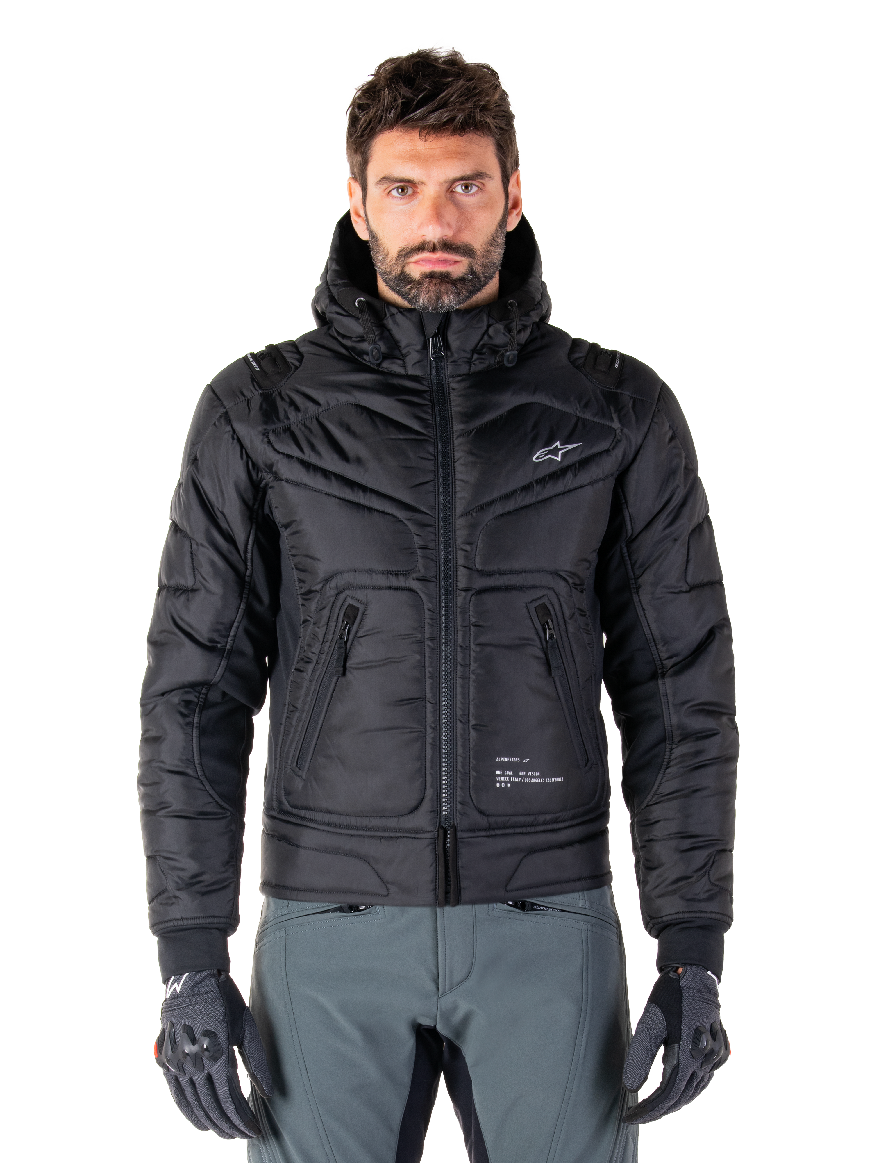 Alpinestars Mohobbs Waterproof Jacket, schwarze gepolsterte Bomber-Motorradjacke, gestepptes Polygewebe mit Frontreißverschluss und Reißverschlusstaschen, kleines weißes Logo auf der Brust, Modell trägt graue Hose und Handschuhe, urbanes Streetwear-Design für Stadtfahrten