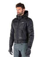 Alpinestars Mohobbs Wasserdichte Jacke, Schwarz, gestepptes wattiertes Bomberjacken-Design mit Kapuze, getragen von einem Mann mit grauer Motorradhose und schwarzen Handschuhen, urbane Streetwear-inspirierte Motorradbekleidung mit einem kleinen weißen Logo auf der Brust.