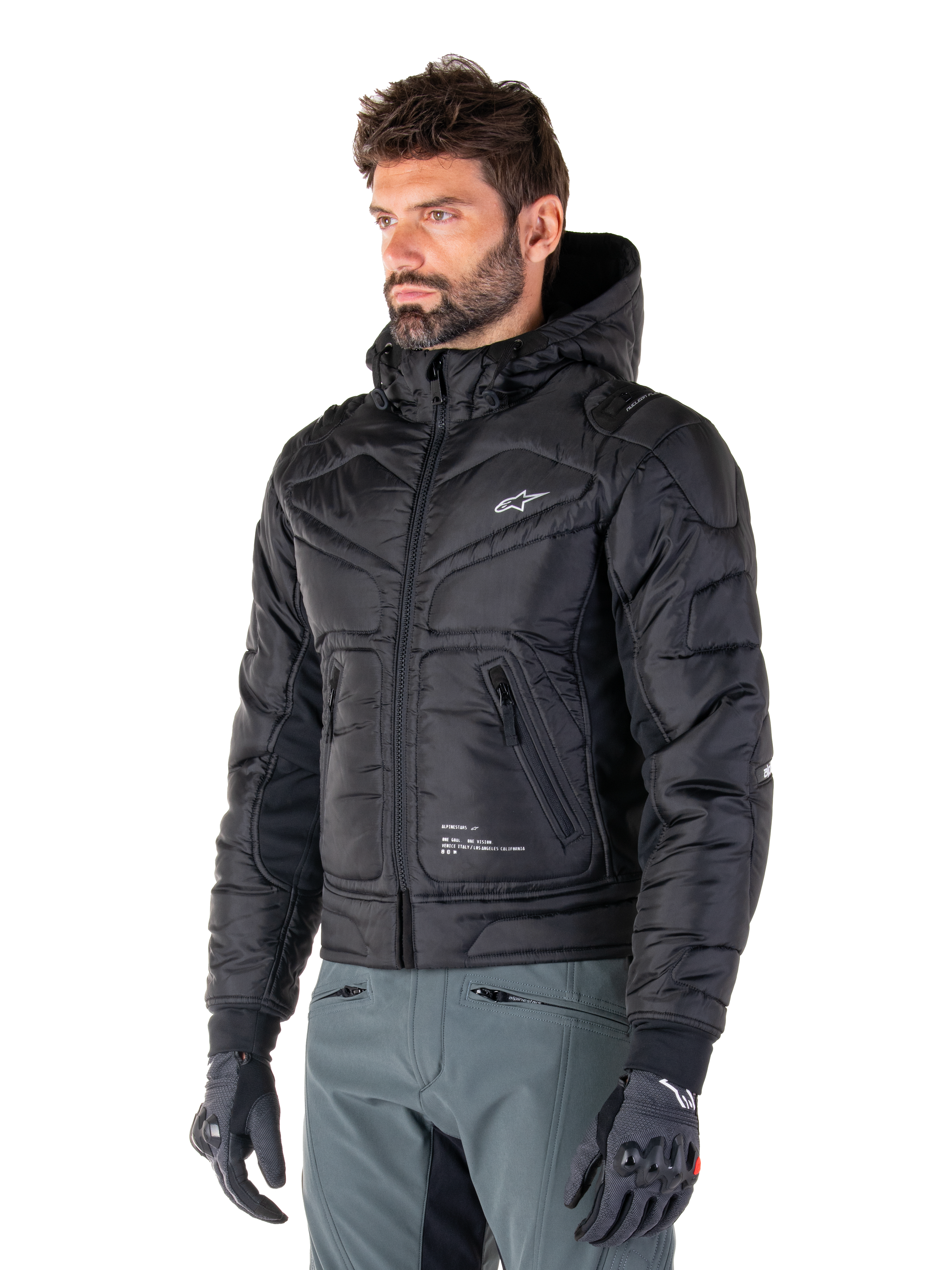 Alpinestars Mohobbs Wasserdichte Jacke, Schwarz, gestepptes wattiertes Bomberjacken-Design mit Kapuze, getragen von einem Mann mit grauer Motorradhose und schwarzen Handschuhen, urbane Streetwear-inspirierte Motorradbekleidung mit einem kleinen weißen Logo auf der Brust.
