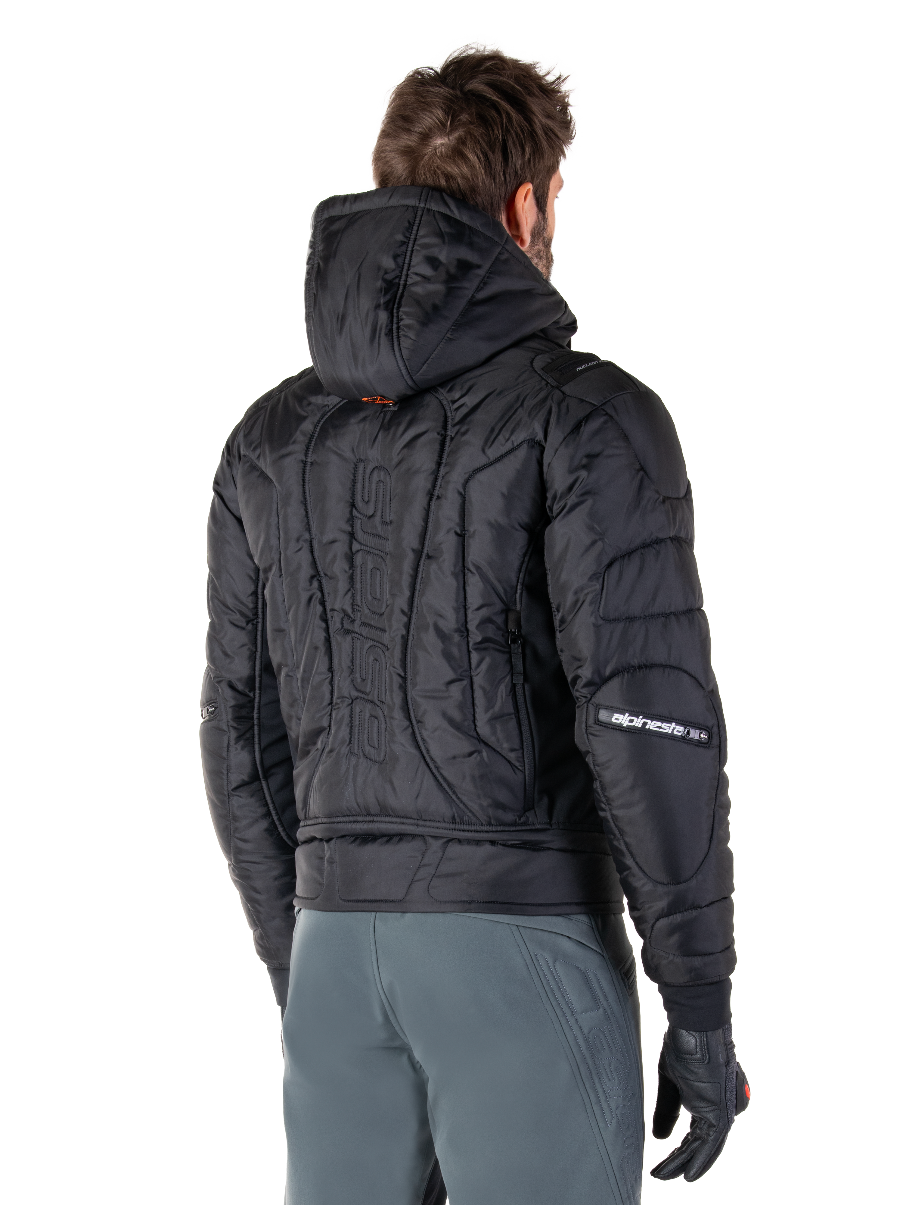Alpinestars Mohobbs Wasserdichte Jacke, Schwarz, Rückansicht mit gesteppter wattierter Konstruktion, Kapuze, vertikaler Astars-Schriftzug auf dem Rücken und Alpinestars-Branding auf den Ärmeln, getragen mit grauer Hose und schwarzen Handschuhen.