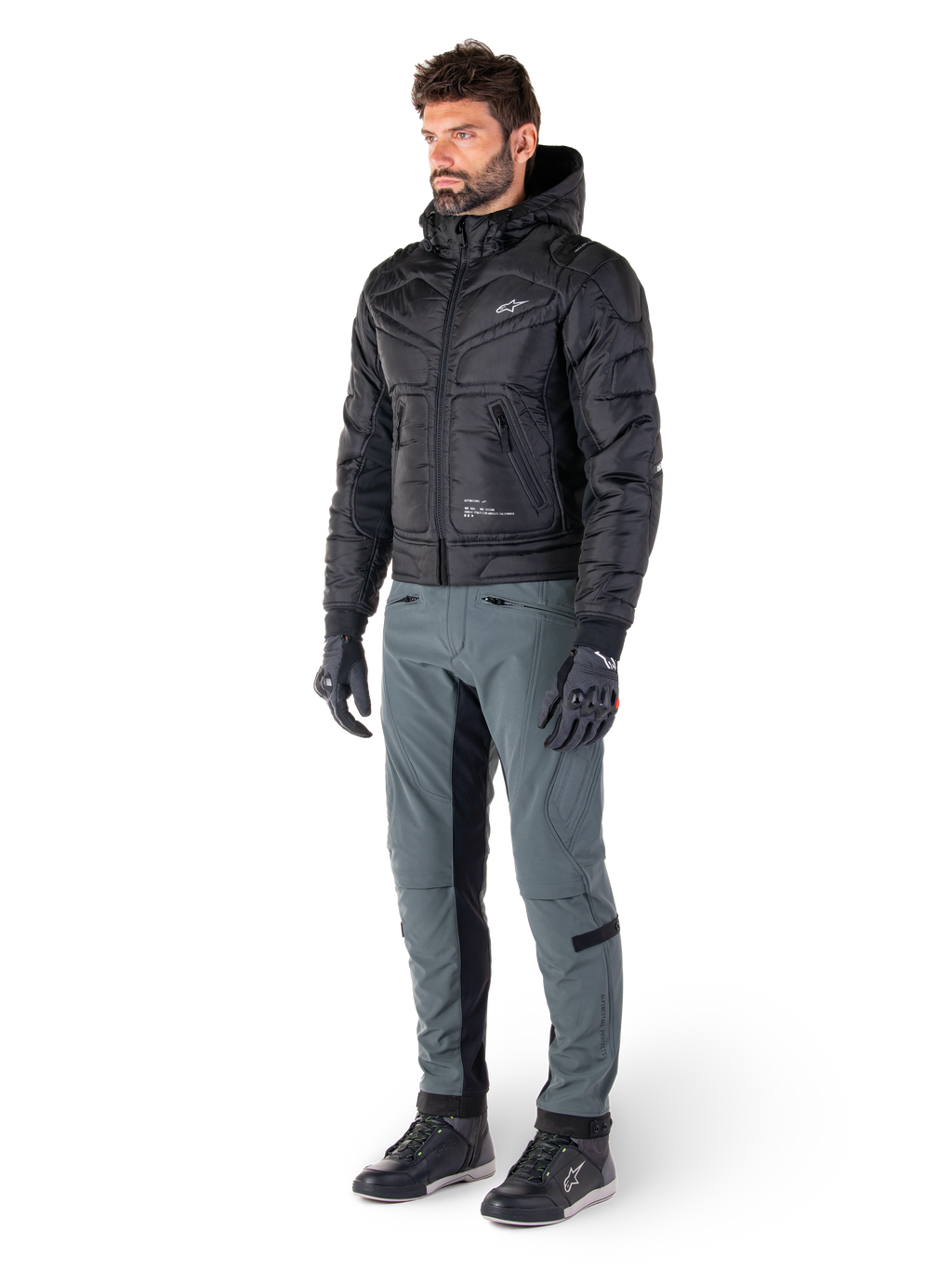 Alpinestars Mohobbs Wasserdichte Jacke, Schwarz, gesteppte urbane Motorrad-Bomberjacke mit Kapuze, getragen von einem Model mit grauer Fahrhose und schwarzen Handschuhen, mit weißem Brustlogo, moderne, von Streetwear inspirierte Schutzausrüstung