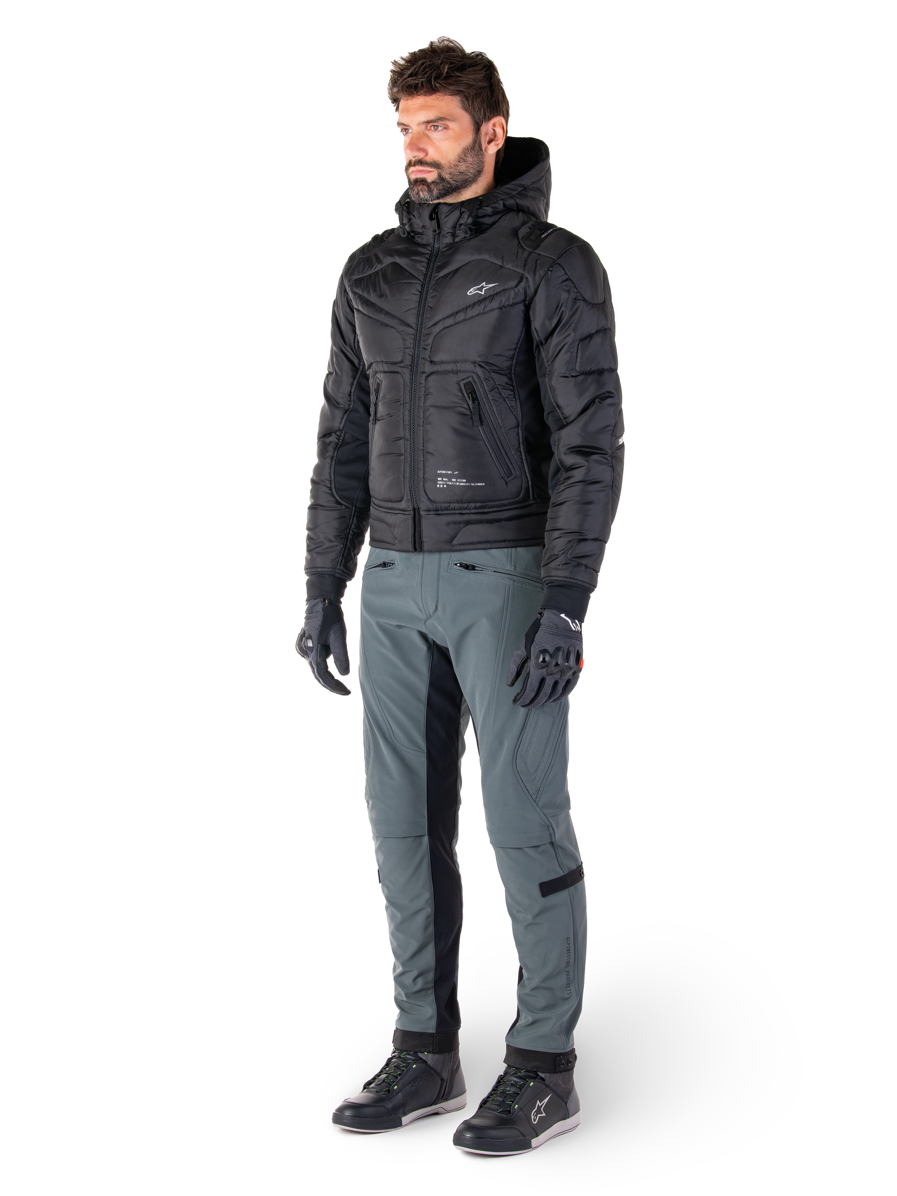 Alpinestars Mohobbs Wasserdichte Jacke, Schwarz, gesteppte urbane Motorrad-Bomberjacke mit Kapuze, getragen von einem Model mit grauer Fahrhose und schwarzen Handschuhen, mit weißem Brustlogo, moderne, von Streetwear inspirierte Schutzausrüstung