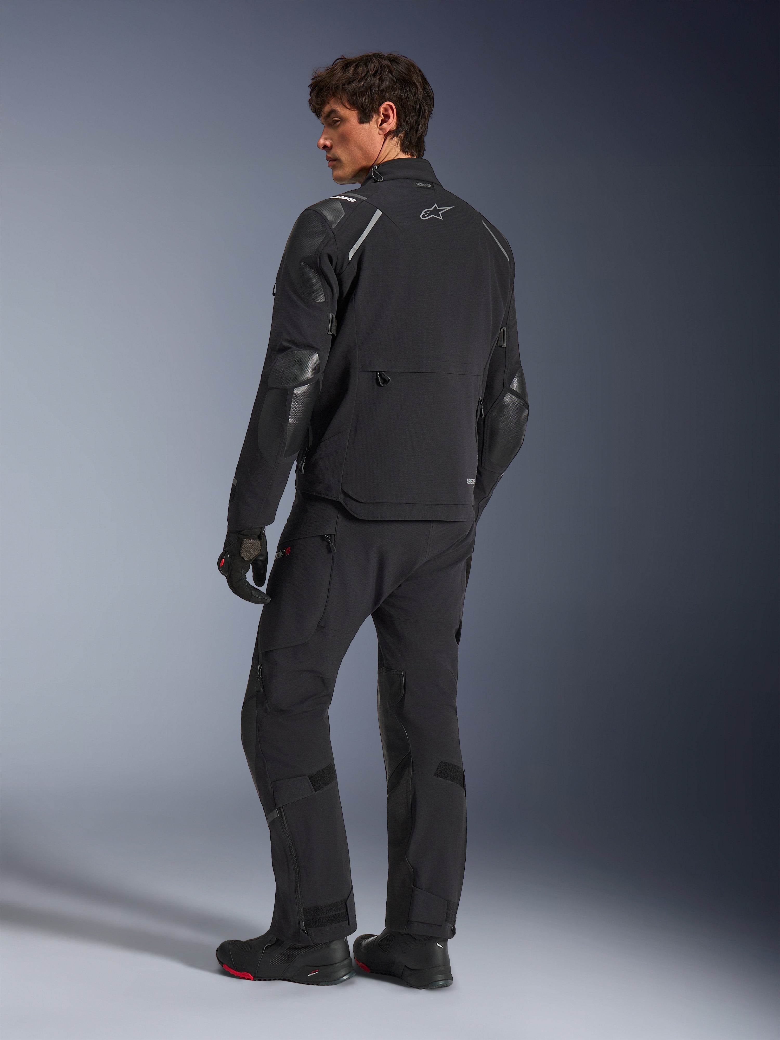 Alpinestars Andes Pro Drystar® XF Jacke, Adventure-Motorradjacke, Schwarz, Rückansicht getragen mit passender Hose und Stiefeln, mit verstärkten Einsätzen und silbernem Stern-Logo, technische Allwetter-Fahrausrüstung