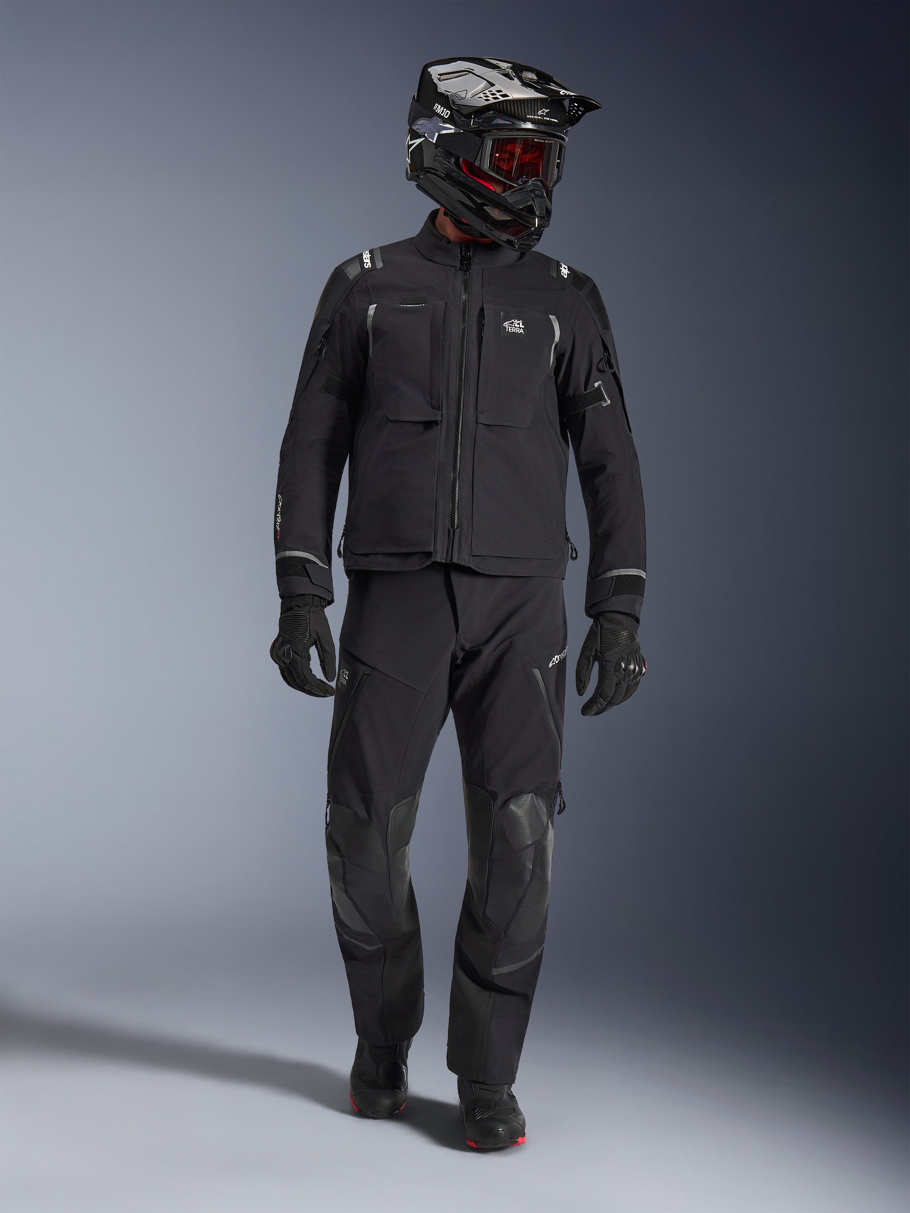 Adventure-Fahrer in der Alpinestars Andes Pro Drystar® XF Jacke in Schwarz und Dunkelgrau, passende schwarze Adventure-Hose, schwarz-weißer Helm mit roter Brille, schwarze Handschuhe und Stiefel, mit großen vorderen Funktionstaschen, reflektierenden Akzenten und technischer 3-Lagen-Laminat-Konstruktion für Allwetter-Touren.