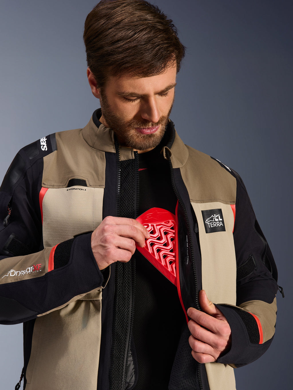 Alpinestars Andes Pro Drystar® XF Jacket in Walnut Aluminum, model demonstrating the red Nucleon chest protector insert into the internal pocket, adventure motorcycle jacket with ALL TERRA branding and Drystar XF logos.