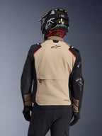 Alpinestars Andes Pro Drystar® XF Jacket in Walnut Aluminum, back view, tan back panel with large cargo pocket, black sleeves with reinforced panels, Alpinestars logo on the upper back, paired with a black helmet and gloves for adventure touring.