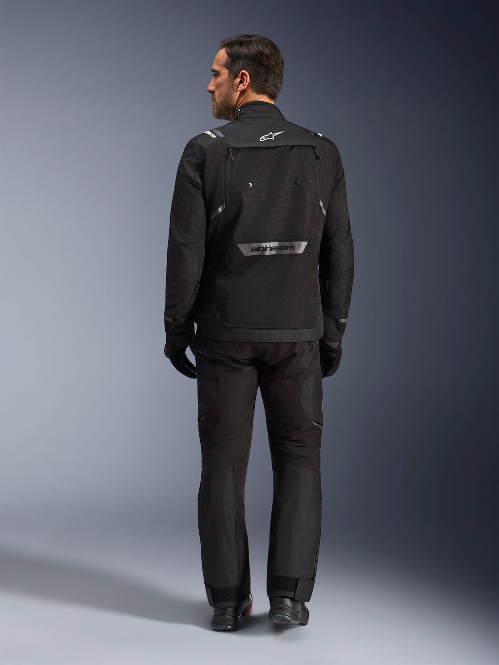 Alpinestars Cusco Drystar® Jacket, Black adventure touring jacket from a rear view, worn with matching black riding pants and gloves, featuring reflective Alpinestars branding on the lower back and durable textile construction for motorcycle road gear.