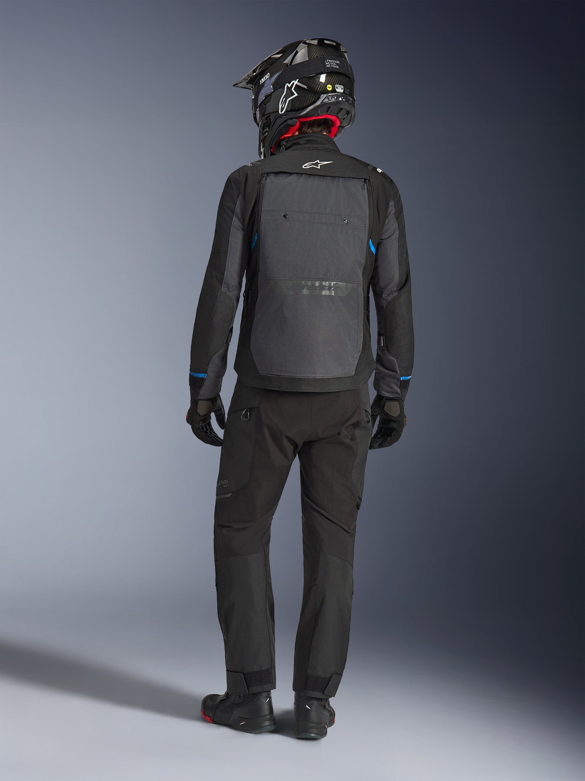 Alpinestars Cusco Drystar® Jacke, Schwarz und Dunkelgrau, Rückansicht eines Fahrers in kompletter Adventure-Touring-Ausrüstung, großes graues Rückenpanel mit Alpinestars-Logo, blaue Ärmelakzente, getragen mit passender schwarzer Hose und Helm.