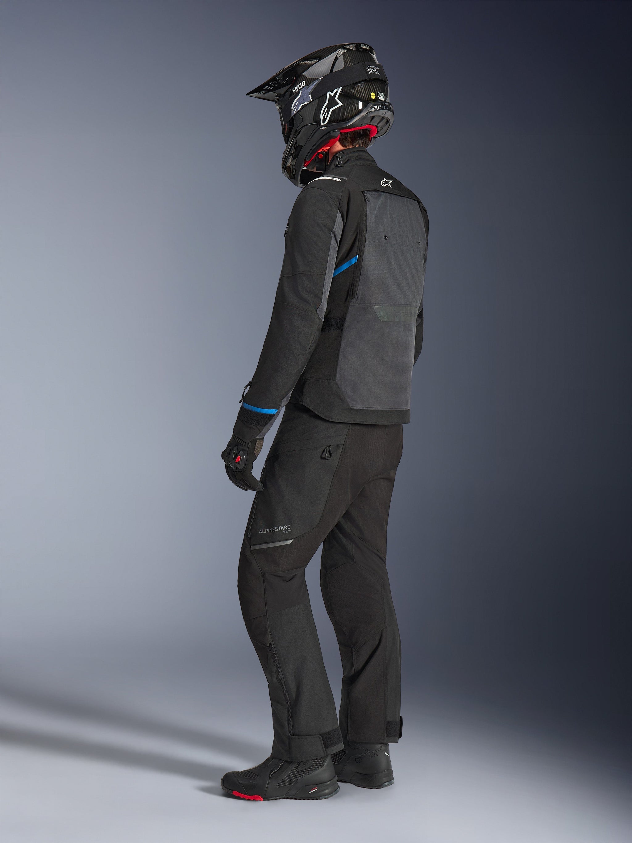 Motorradfahrer in der Alpinestars Cusco Drystar® Jacke in Schwarz und Dunkelgrau mit blauen Akzenten, kombiniert mit einer schwarzen Motorradhose, einem SM10 Carbon-Helm und schwarzen Handschuhen, Rückansicht mit Fokus auf das große Belüftungspanel und das Adventure-Touring-Design.