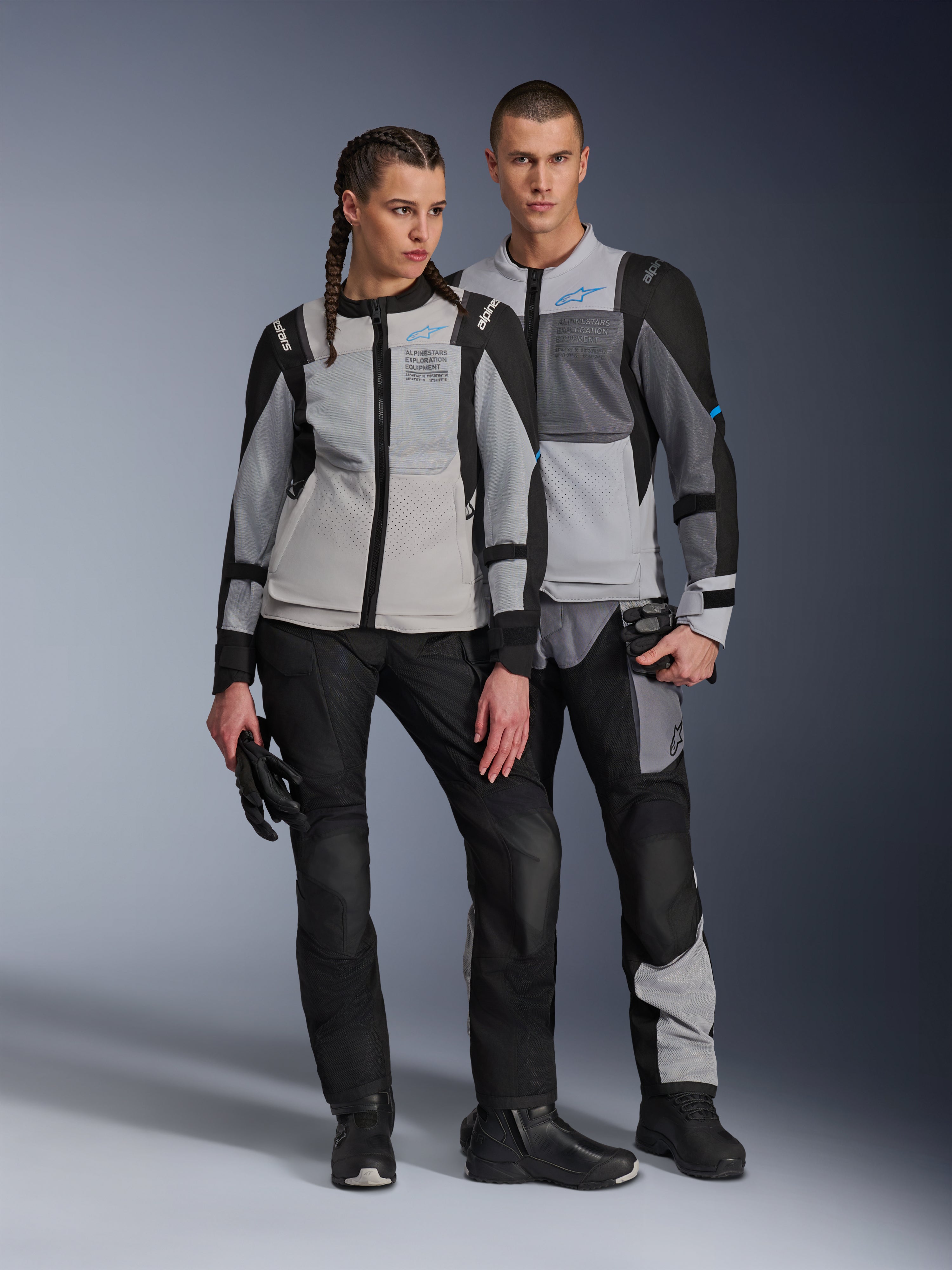 Male and female models wearing Alpinestars ST-2 Air Jackets in Ice Gray Dark Gray and Black, technical summer touring motorcycle gear featuring large mesh panels and exploration equipment branding, paired with black riding pants, gloves, and boots.