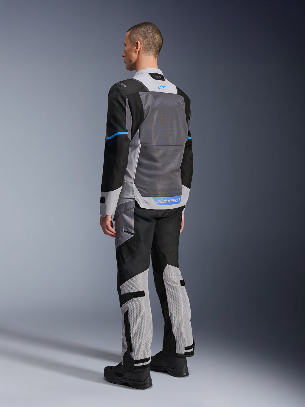 Male model from behind wearing the Alpinestars ST-2 Air Jacket in Ice Gray, Dark Gray, and Black, featuring a large grey mesh back panel, blue logo accents, and matching motorcycle pants for summer touring.