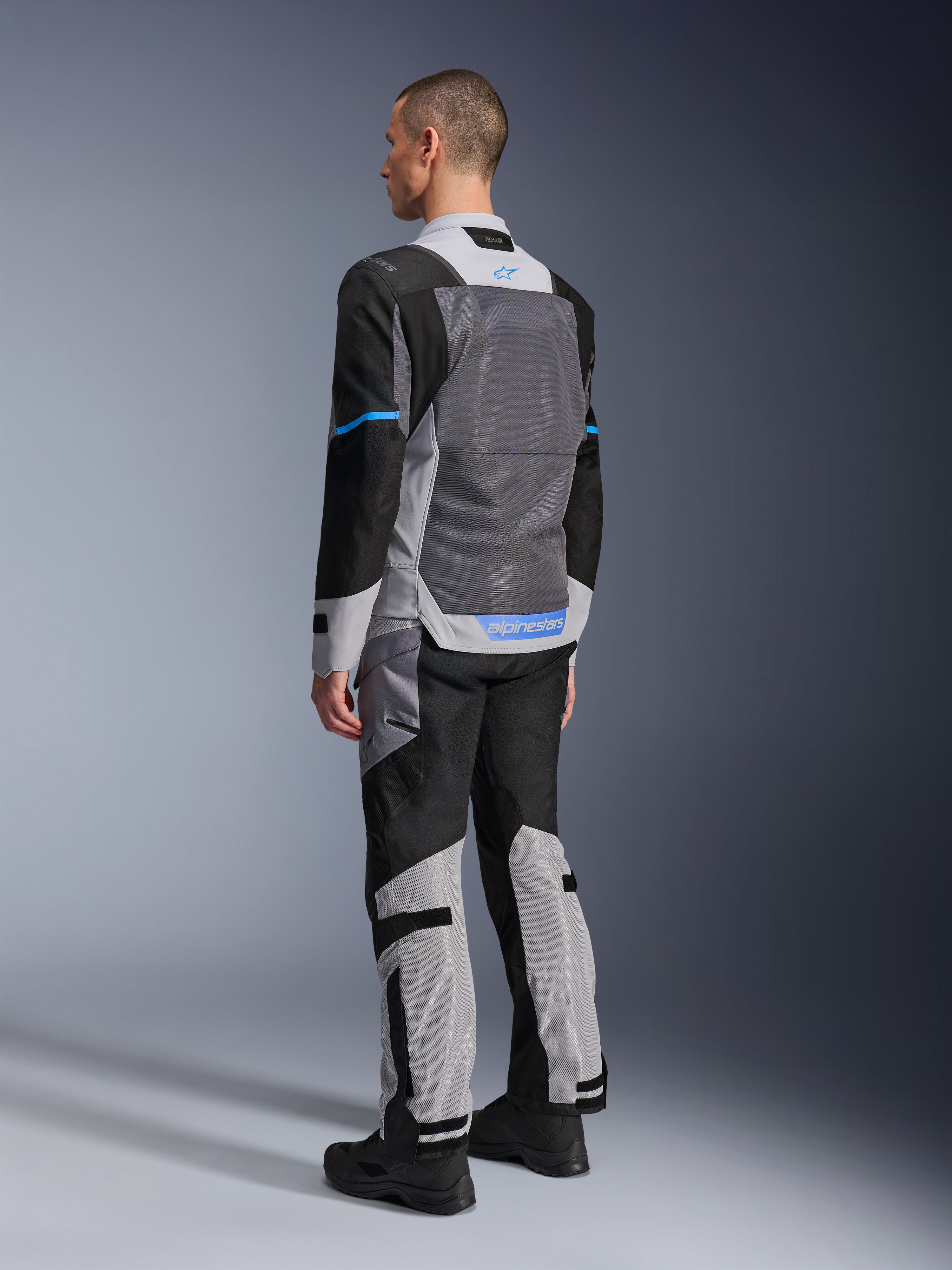 Male model from behind wearing the Alpinestars ST-2 Air Jacket in Ice Gray, Dark Gray, and Black, featuring a large grey mesh back panel, blue logo accents, and matching motorcycle pants for summer touring.