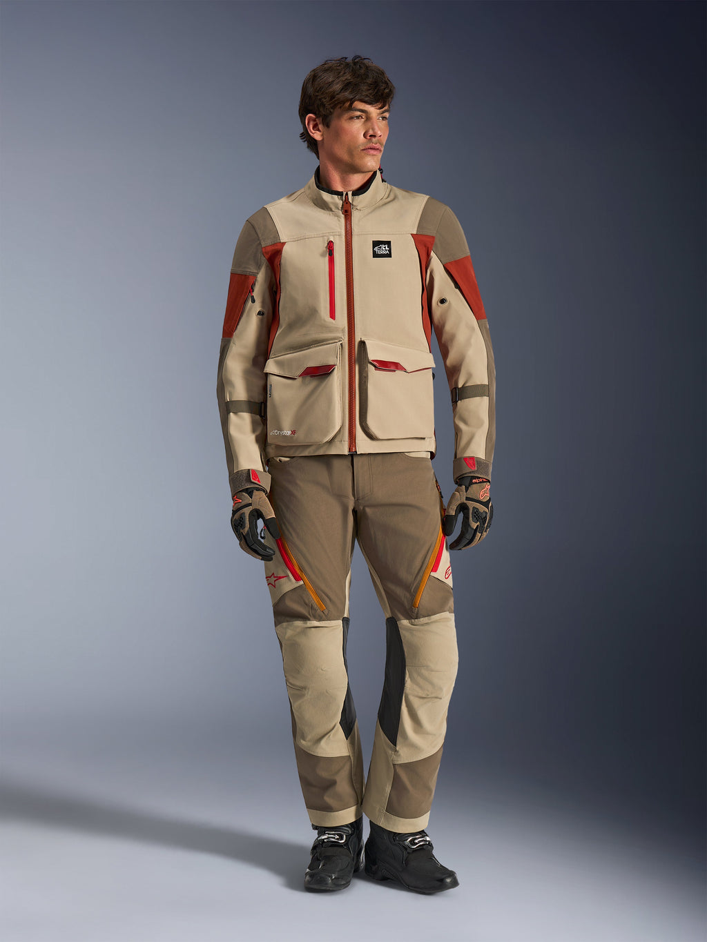 Maxdura Drystar®XF Laminated Armored Shell, Adventure-Motorradjacke in Aluminum Walnut Rust Brown, getragen von einem männlichen Model mit passender Adventure-Hose, Handschuhen und Stiefeln, Ganzkörper-Studioaufnahme einer professionellen Tourenausrüstung.