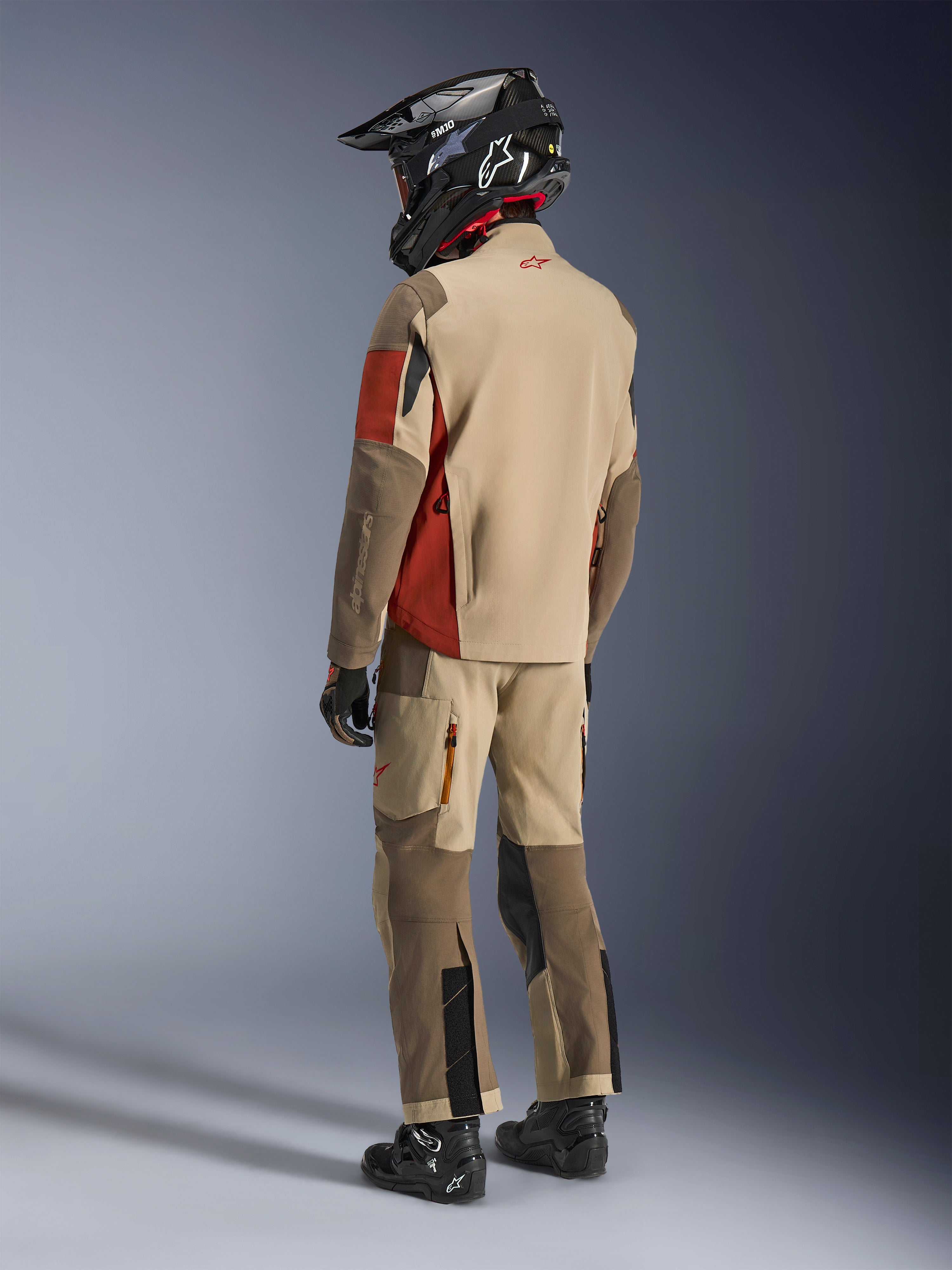 Adventure-Fahrer in der Rückansicht mit der Alpinestars Maxdura Drystar®XF Laminated Armored Shell in Aluminum Walnut Rust Brown, kombiniert mit passender Motorradhose, schwarzem Offroad-Helm und schwarzen Stiefeln, komplettes technisches Touren-Outfit.