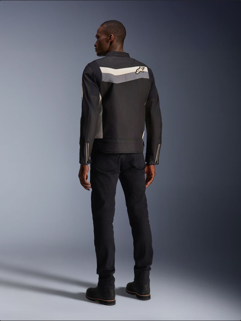 Männliches Model von hinten in der Alpinestars T-Dyno WR Jacke in Black Dark Gray Ecru, Textil-Motorradjacke mit horizontalem Streifen und Stern-Logo auf dem Rücken, kombiniert mit schwarzer Motorradhose und Stiefeln, urbane Motorradausrüstungs-Ästhetik.
