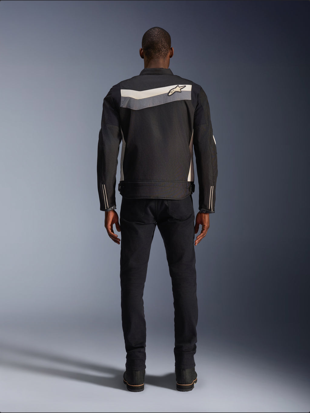 Alpinestars T-Dyno WR Jacke, Textil-Motorradjacke, Schwarz, Dunkelgrau und Ecru, Rückansicht mit horizontalem Streifen über den Schultern und kleinem Stern-Logo, getragen von einem Model mit schwarzer Hose und Stiefeln, Vintage-inspirierte Motorradausrüstung