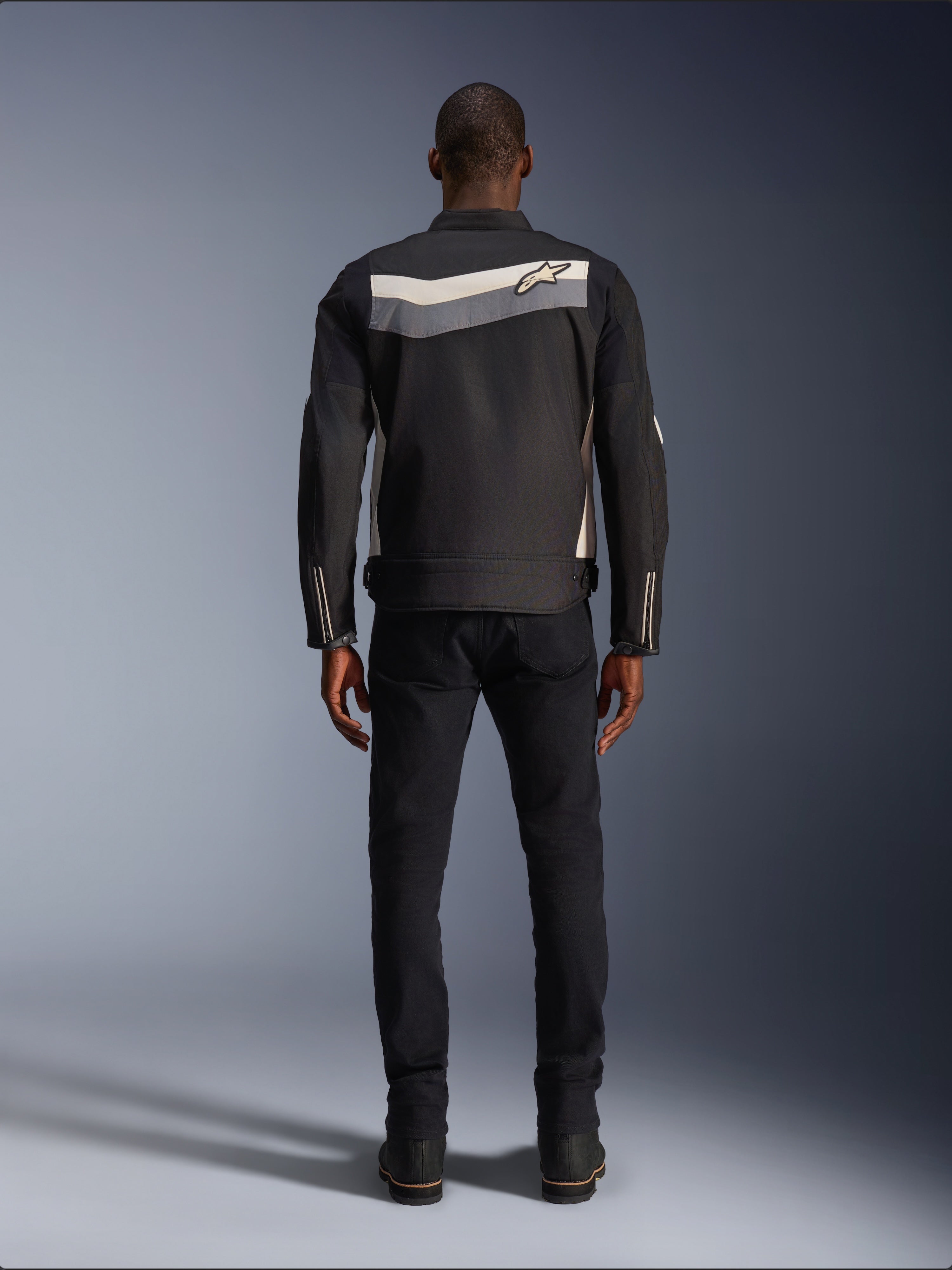 Alpinestars T-Dyno WR Jacke, Textil-Motorradjacke, Schwarz, Dunkelgrau und Ecru, Rückansicht mit horizontalem Streifen über den Schultern und kleinem Stern-Logo, getragen von einem Model mit schwarzer Hose und Stiefeln, Vintage-inspirierte Motorradausrüstung