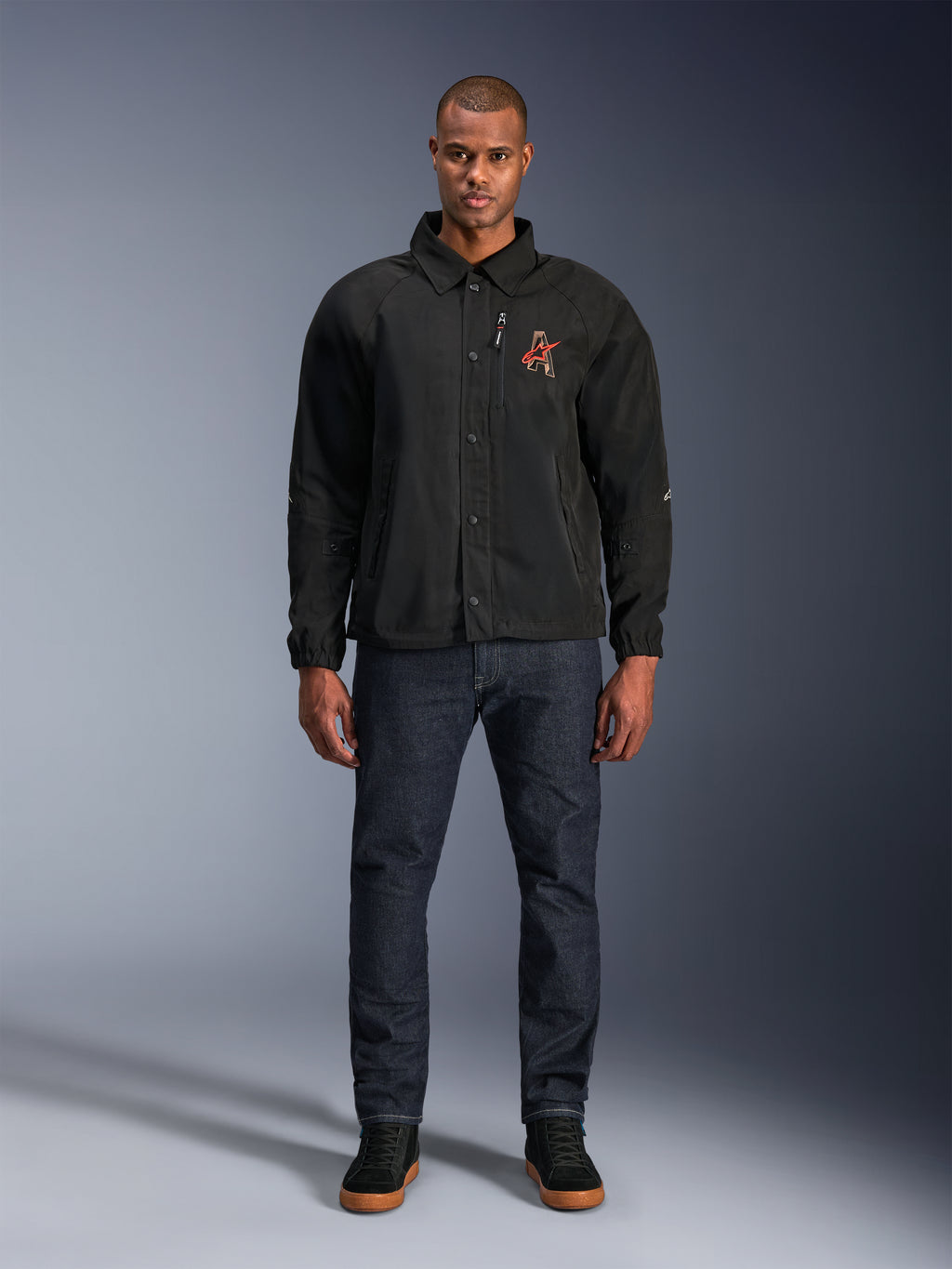 Alpinestars Revved WR Jacke in Schwarz, getragen von einem Modell mit dunkelblauen Jeans und schwarzen Sneakern, Vorderansicht, mit Druckknopfverschluss und einem 3D-Stickerei-Logo auf der Brust, moderne Urban-Motorradausrüstung.