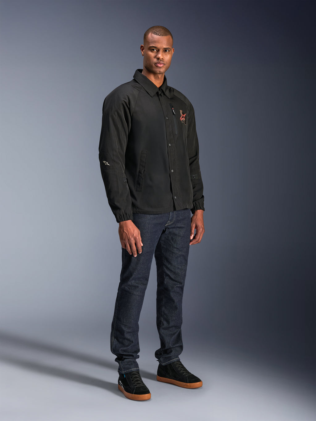 Alpinestars Revved WR Jacke in Schwarz, Motorradjacke im urbanen Coach-Stil, getragen von einem Model mit dunkelblauen Jeans und schwarzen Sneakern, mit Druckknopfleiste vorne und gesticktem Logo, minimalistisches und sportliches Design für Straßenfahrten