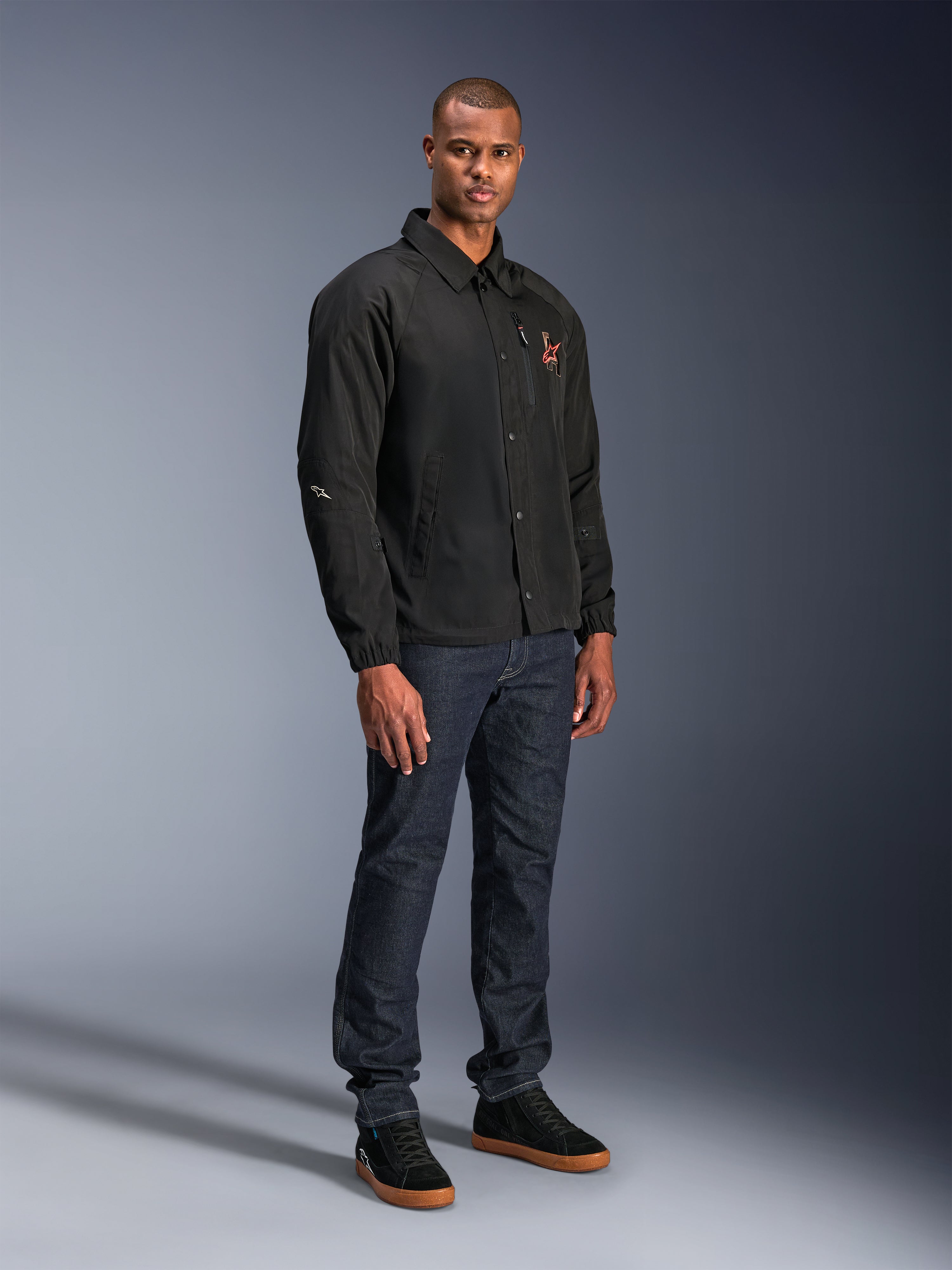 Alpinestars Revved WR Jacke in Schwarz, Motorradjacke im urbanen Coach-Stil, getragen von einem Model mit dunkelblauen Jeans und schwarzen Sneakern, mit Druckknopfleiste vorne und gesticktem Logo, minimalistisches und sportliches Design für Straßenfahrten