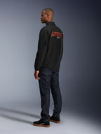 Alpinestars Revved WR Jacke, Schwarz, mit großem orangefarbenem 3D-gesticktem Alpinestars-Branding auf dem Rücken, Dreiviertel-Rückansicht eines Models in dunkelblauen Jeans und schwarzen Sneakern, moderne urbane Motorradjacke mit Coach-Jacken-Silhouette