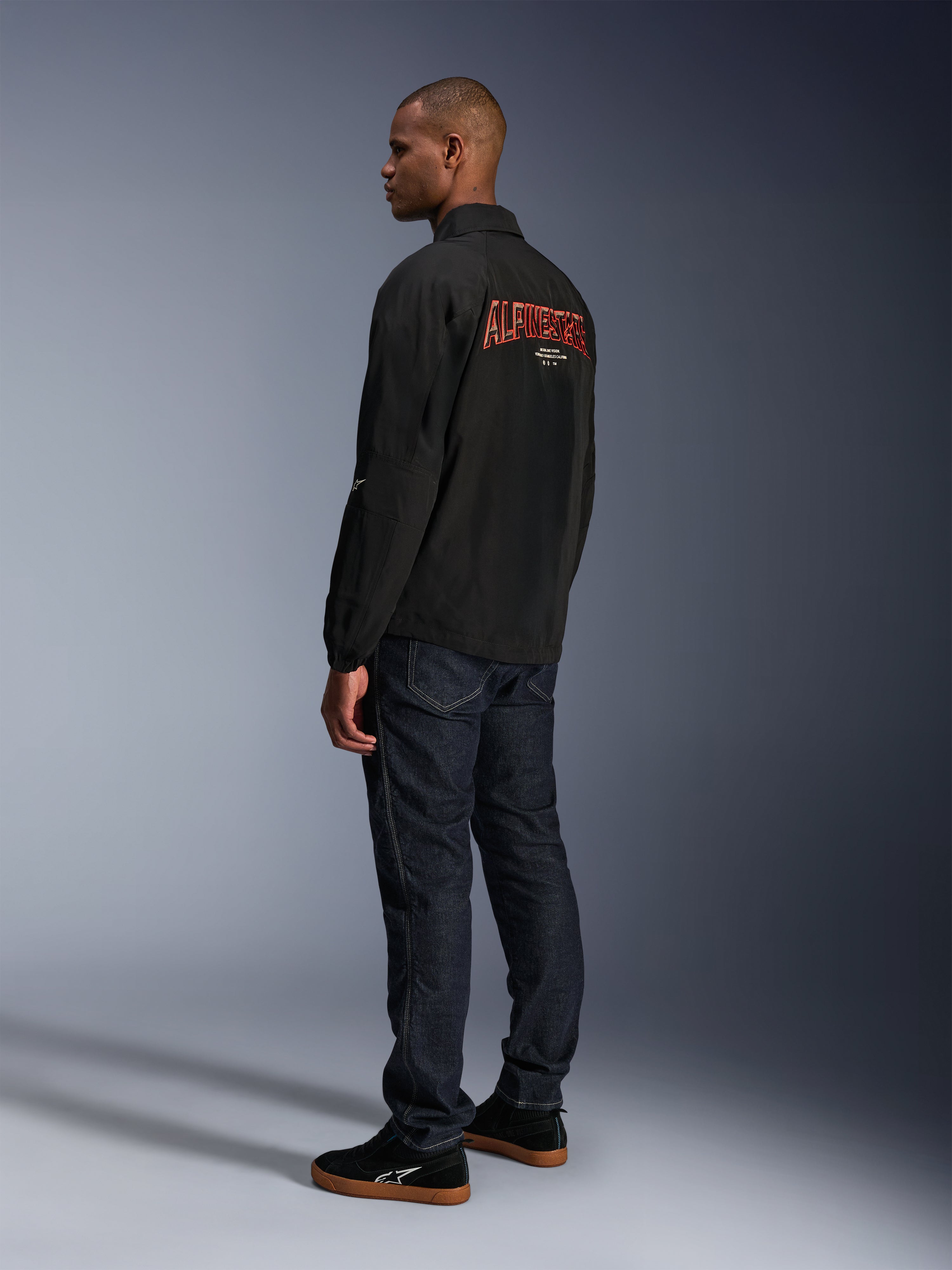 Alpinestars Revved WR Jacke, Schwarz, mit großem orangefarbenem 3D-gesticktem Alpinestars-Branding auf dem Rücken, Dreiviertel-Rückansicht eines Models in dunkelblauen Jeans und schwarzen Sneakern, moderne urbane Motorradjacke mit Coach-Jacken-Silhouette