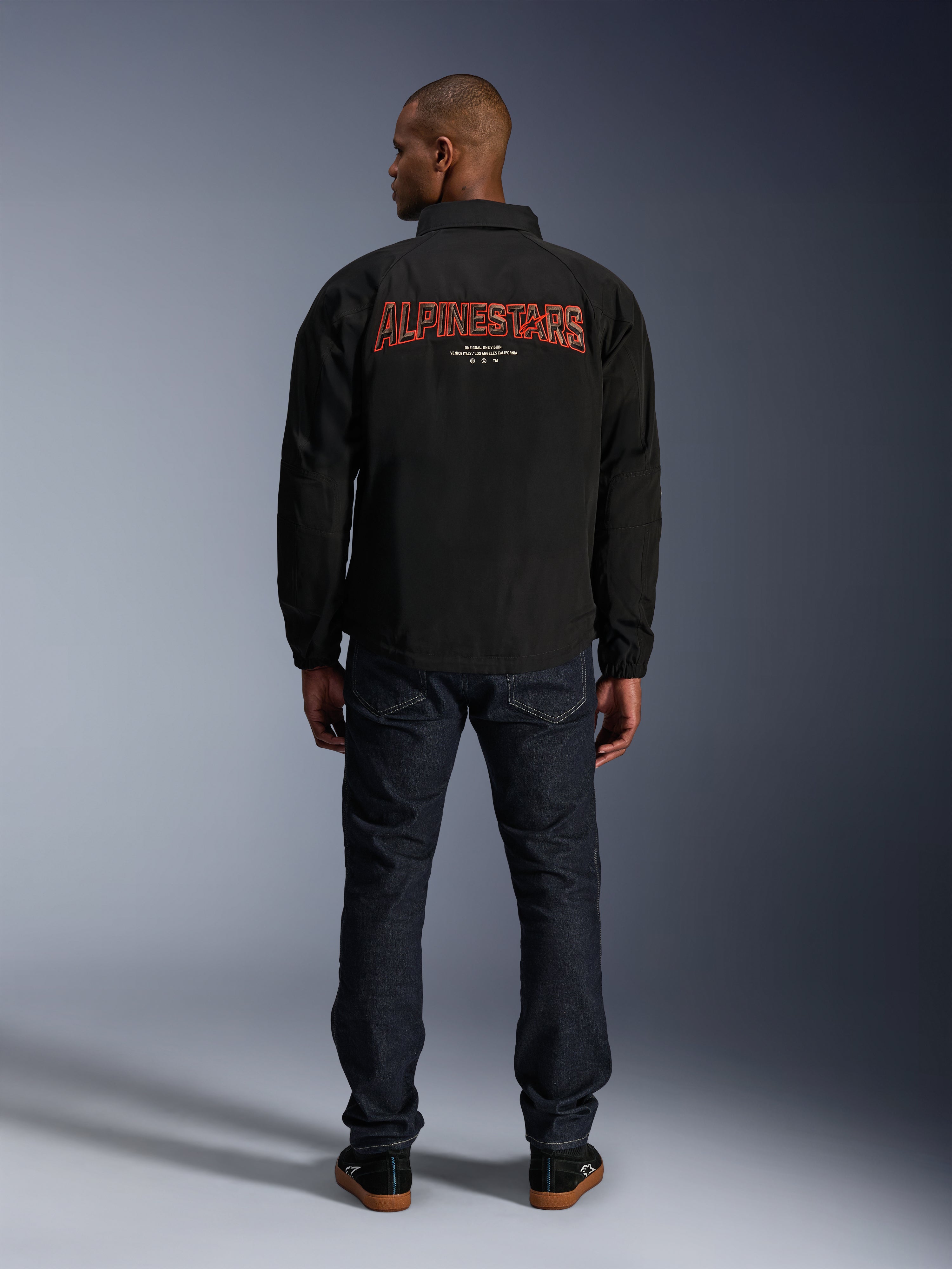 Alpinestars Revved WR Jacke, Schwarz, Rückansicht mit großer roter 'ALPINESTARS' Stickerei im 3D-Effekt, urbane Jacke im Coach-Stil, getragen mit dunkler Denim-Jeans und schwarzen Sneakern, minimalistische Motorradbekleidung