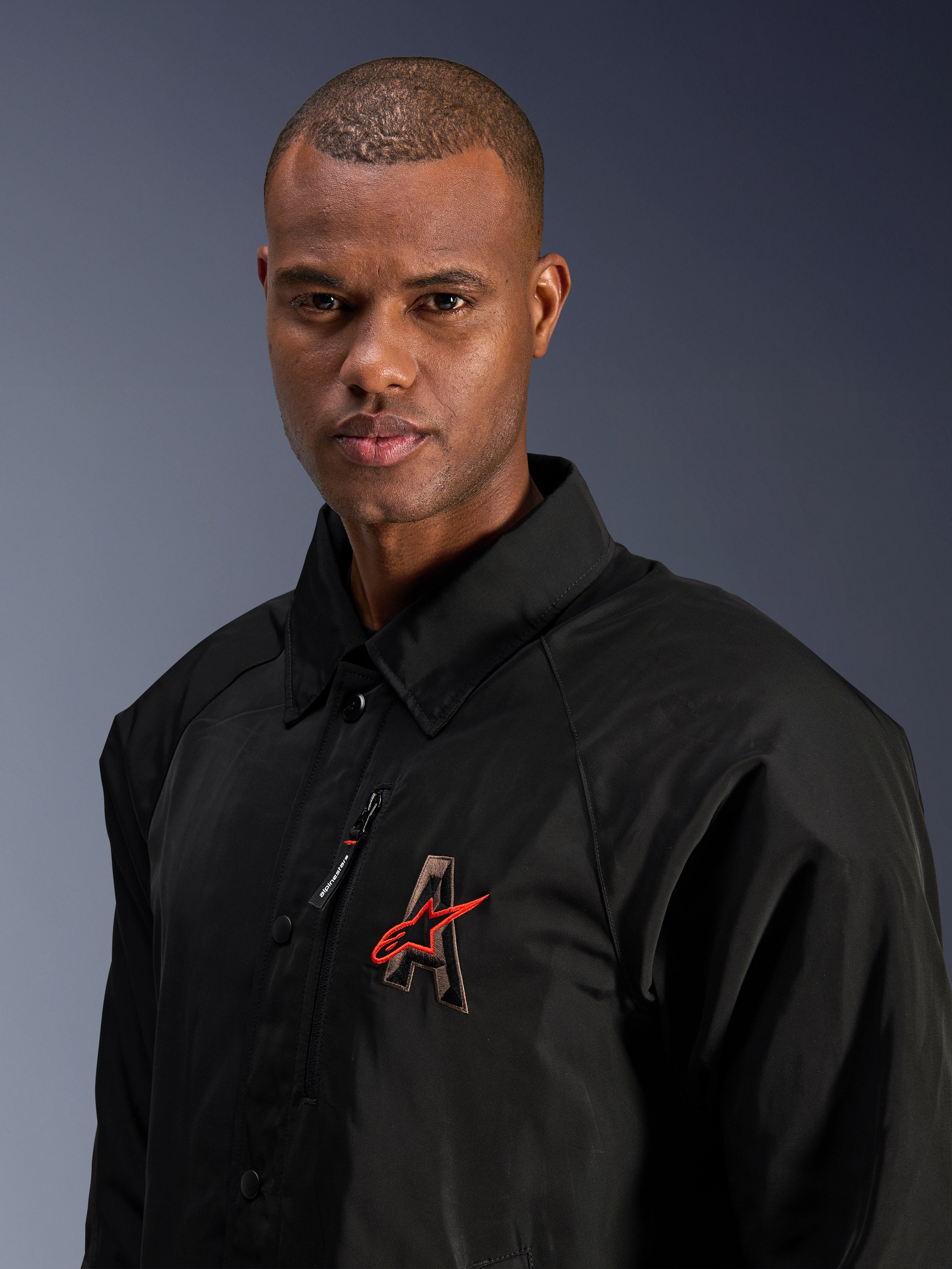 Alpinestars Revved WR Jacke, Schwarz, Nahaufnahme eines Mannes in der wasserabweisenden Satin-Motorradjacke, mit 3D-gesticktem „A“-Logo und rotem Alpinestars-Stern, Brusttasche mit Reißverschluss und Kragendesign für einen urbanen, zeitgenössischen Fahrstil.