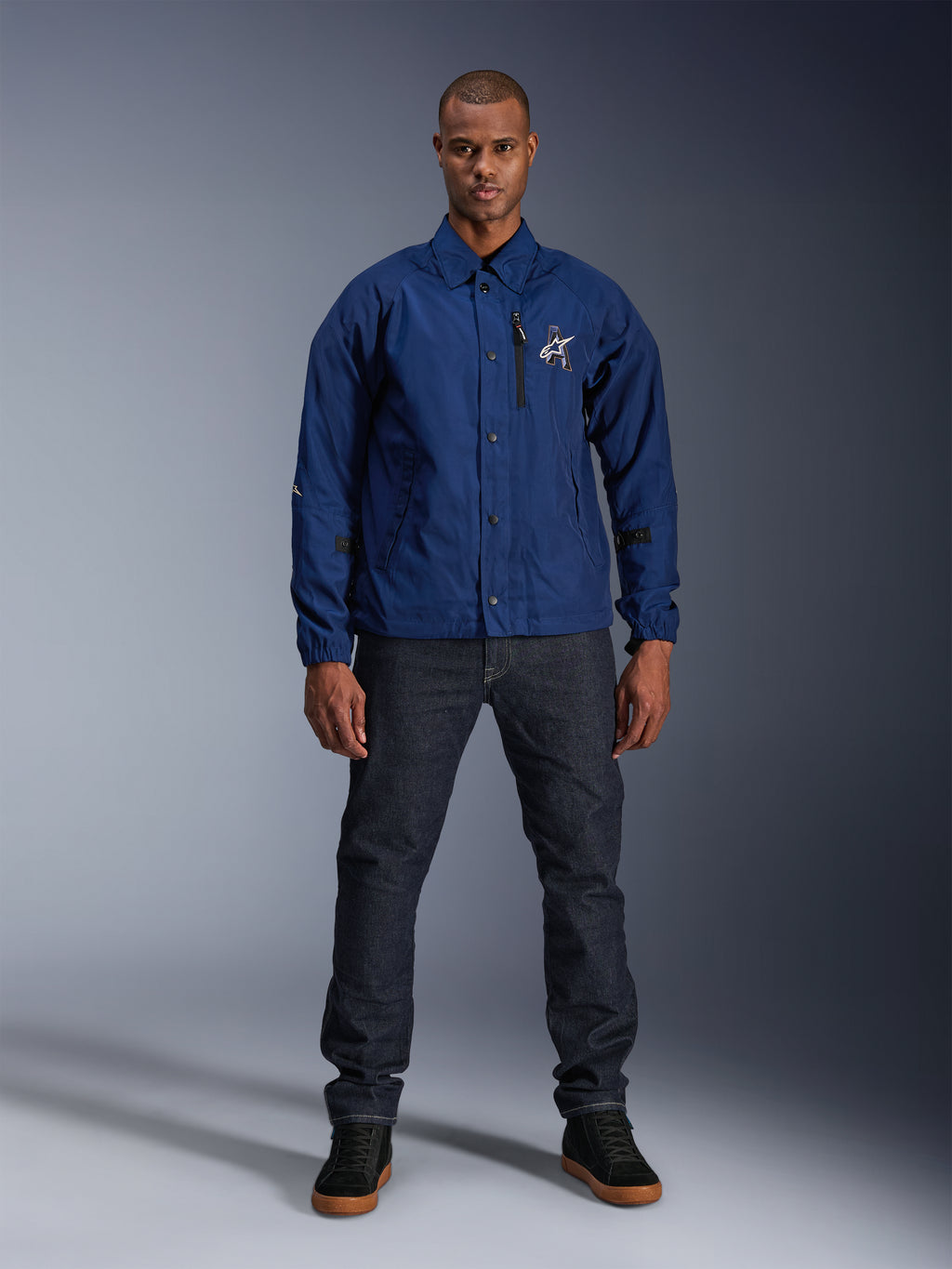 Alpinestars Revved WR Jacke, Marineblau, Motorradjacke im Coach-Stil, getragen von einem Mann mit dunklen Denim-Jeans und schwarzen Sneakern, mit Druckknopfverschluss und 3D-Alpinestars-Logo, urbane moderne Streetwear