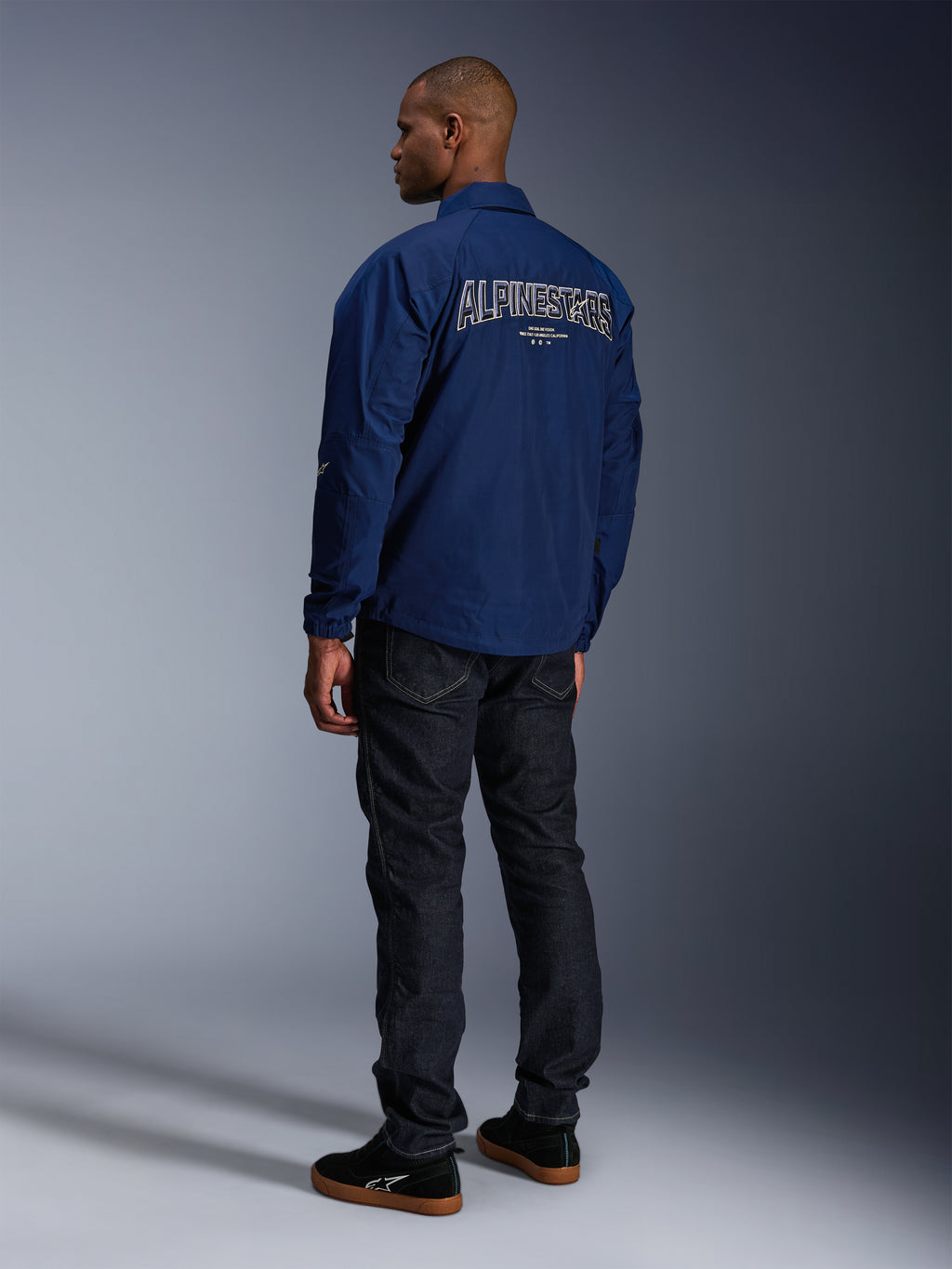 Model in der Alpinestars Revved WR Jacke in Marineblau von hinten, mit großer weiß umrandeter „ALPINESTARS“ Stickerei auf dem Rücken, kombiniert mit einer dunklen Jeanshose und schwarzen Sneakern, urbane Motorrad-Ästhetik.