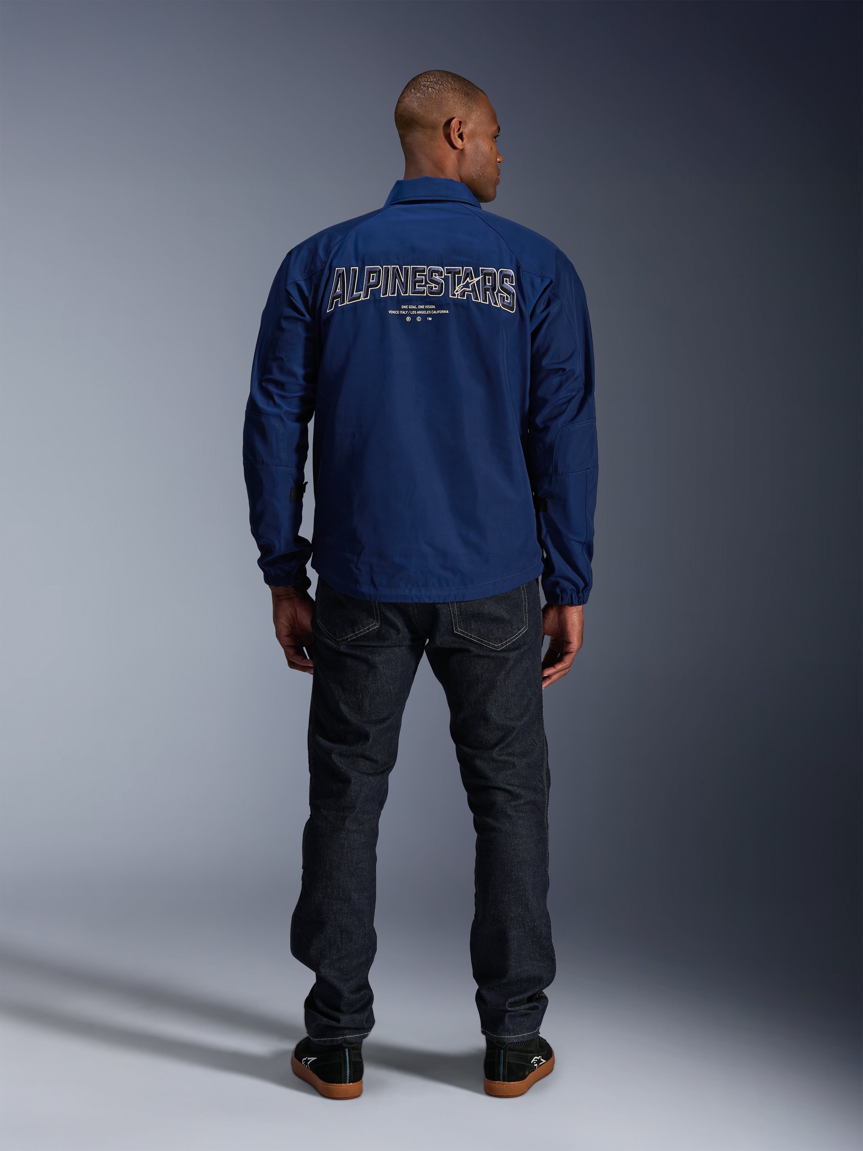 Alpinestars Revved WR Jacke in Marineblau, Rückansicht, mit großer „ALPINESTARS“-Stickerei in 3D-Optik auf dem Rücken, kombiniert mit dunklen Denim-Jeans und schwarzen Sneakern, urbane Motorradbekleidung.