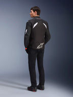 Male model from the back wearing the Alpinestars T-GP R V3 Drystar® Jacket in Black and White, featuring white shoulder accents and a large white logo on the lower back, paired with black riding pants and boots, sport textile motorcycle jacket.