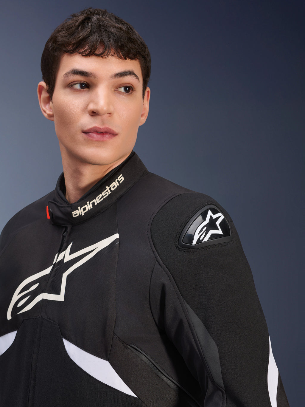 Alpinestars T-GP R V3 Drystar® Jacket, Black and White, worn by a male model, featuring white Alpinestars logos on the collar and chest, black DFS shoulder slider with a white star, detailed lifestyle shot of a waterproof sport textile jacket