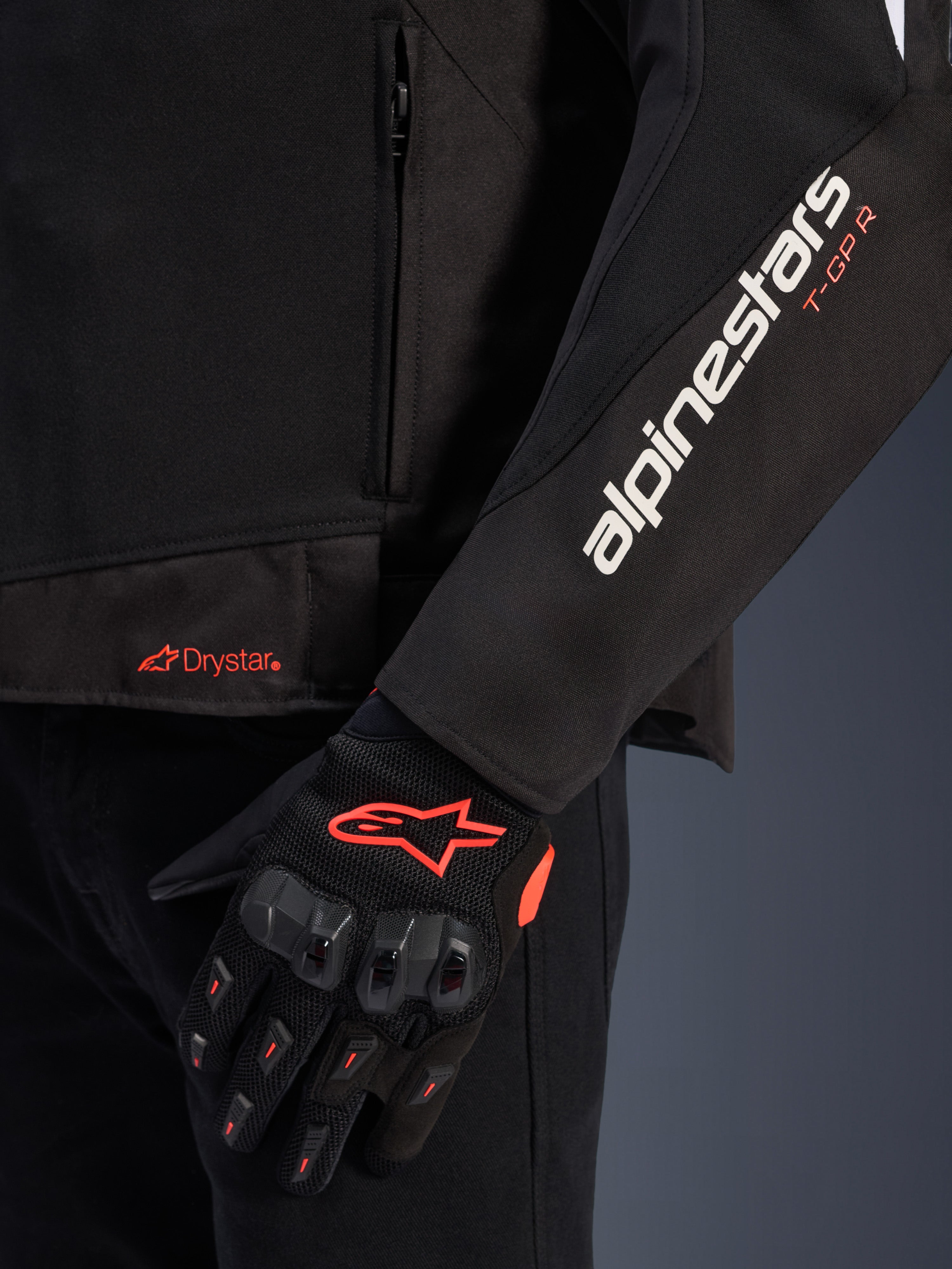 Alpinestars T-GP R V3 Drystar® Jacket, black textile motorcycle jacket, close-up view showing white alpinestars and orange T-GP R sleeve branding, orange Drystar logo on the hem, paired with black protective mesh gloves with red accents.
