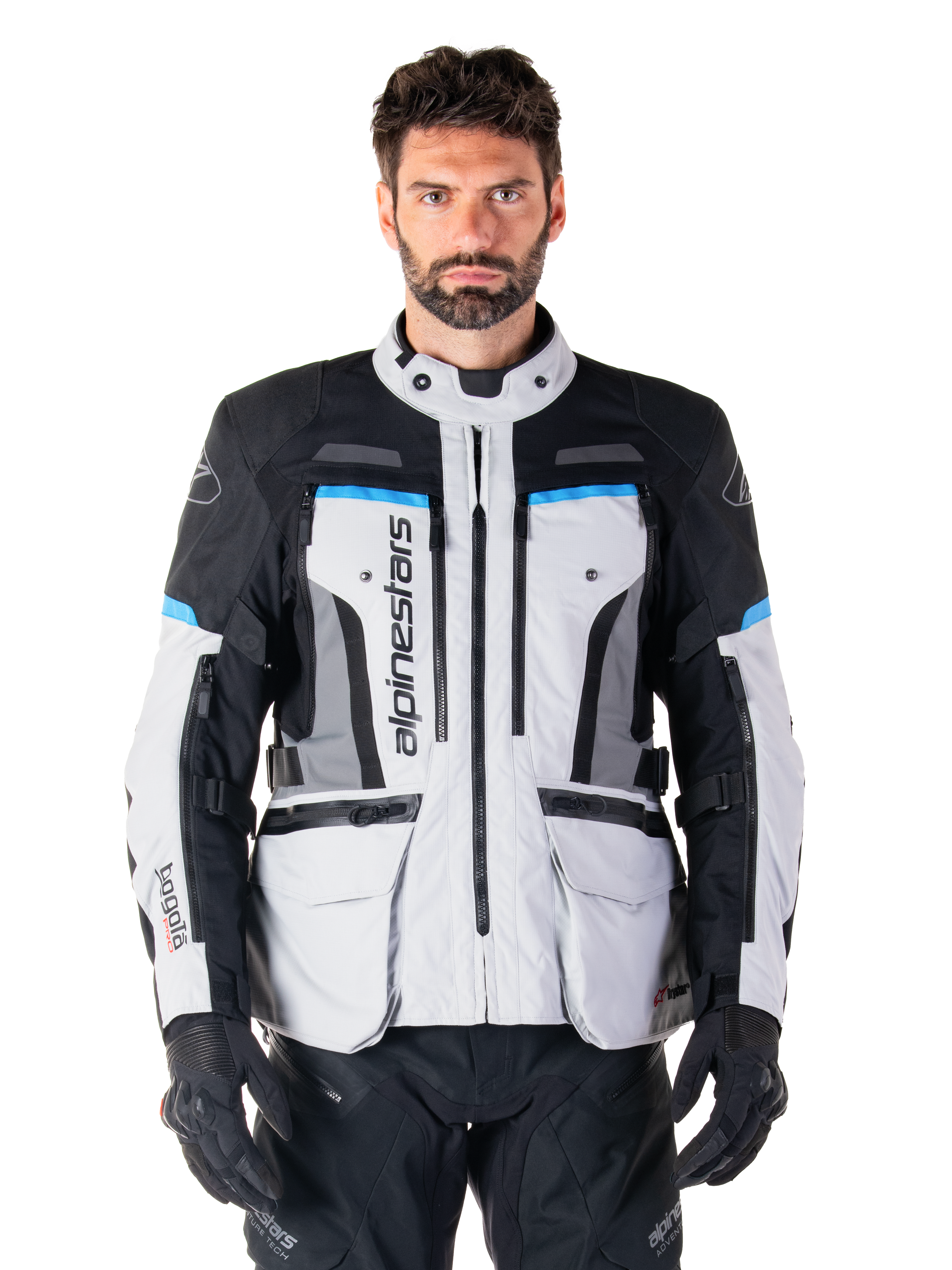 Alpinestars Bogota Pro Drystar Jacke, Motorrad-Adventure-Touringjacke, Eisgrau mit schwarzen und cyanblauen Akzenten, Vorderansicht mit Belüftungspanelen und Cargo-Taschen, getragen von einem Modell mit schwarzer Motorradhose und Handschuhen