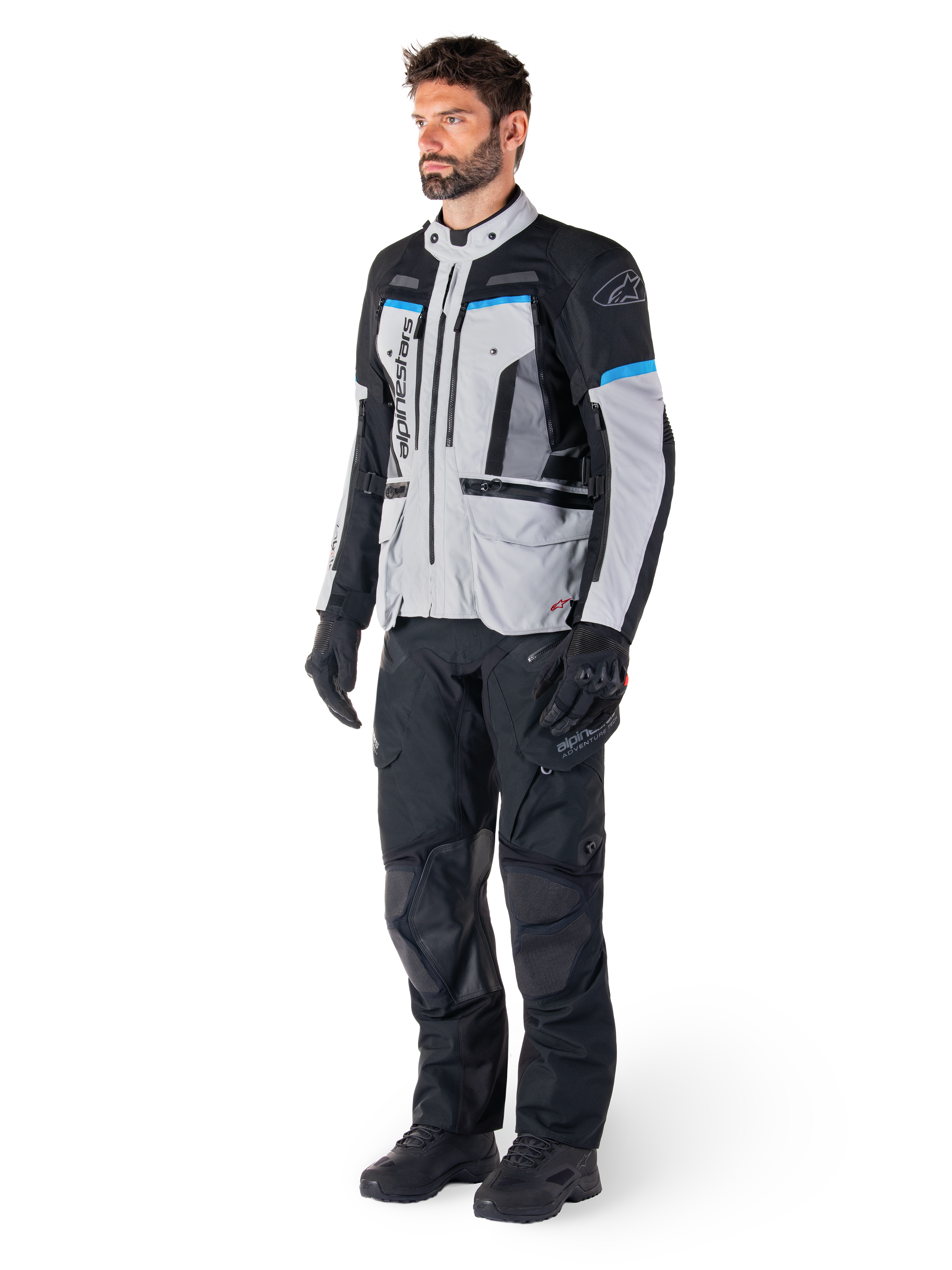 Männlicher Motorradfahrer in der Alpinestars Bogotá Pro Drystar® Jacke in Eisgrau, Schwarz und Cyan, kombiniert mit schwarzer technischer Motorradhose und Handschuhen, Adventure-Touring-Ausrüstung für Performance bei jedem Wetter.