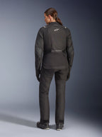 Motorradfahrerin in der Rückansicht mit der Alpinestars Stella Andes V4 Drystar® Jacke, Schwarz, kombiniert mit passender Motorradhose, weißes Astar-Logo auf dem Rücken, verlängerter Rückensaum, komplettes Adventure-Touring-Fahroutfit inklusive schwarzer Handschuhe und Stiefel