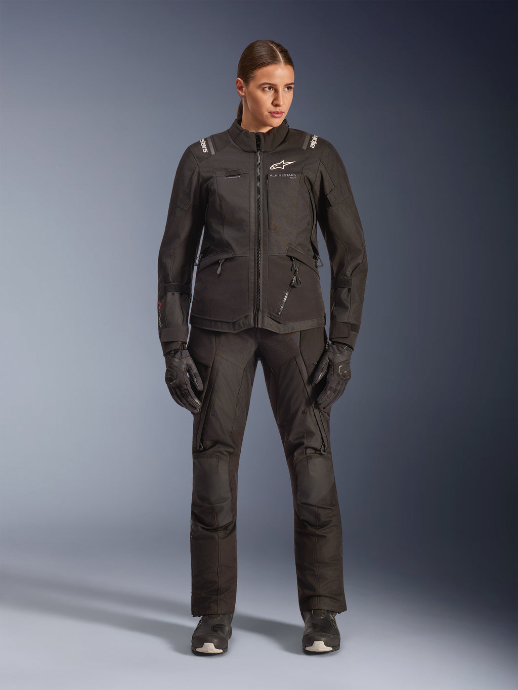 Motorradfahrerin mit Alpinestars Stella Andes V4 Drystar® Jacke in Schwarz, kombiniert mit passender schwarzer Adventure-Tourenhose und Handschuhen, technische Textilkonstruktion mit mehreren Funktionstaschen und weißem Alpinestars-Branding, komplette Allwetter-Motorradausrüstung