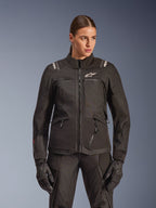 Alpinestars Stella Andes V4 Drystar Jacke, Damen-Motorrad-Tourenjacke, Schwarz, getragen von einem Modell mit passender Hose und Handschuhen, mit Reißverschluss vorne, weißem Logo auf der Brust und Alpinestars-Branding auf den Schultern, elegante Allwetterausrüstung.