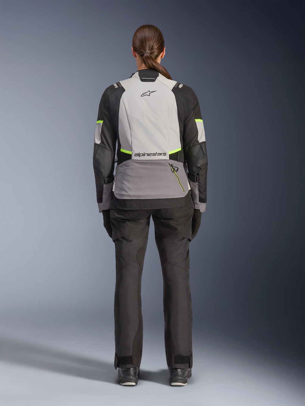 Alpinestars Stella Andes V4 Drystar® Jacke für Damen, Fahrerin in der Rückansicht, Eisgrau, Dunkelgrau und Schwarz mit gelben Fluo-Akzenten, mit einer großen Rückentasche und Alpinestars-Logo, kombiniert mit passender schwarzer Adventure-Hose, Allwetter-Motorrad-Tourenausrüstung