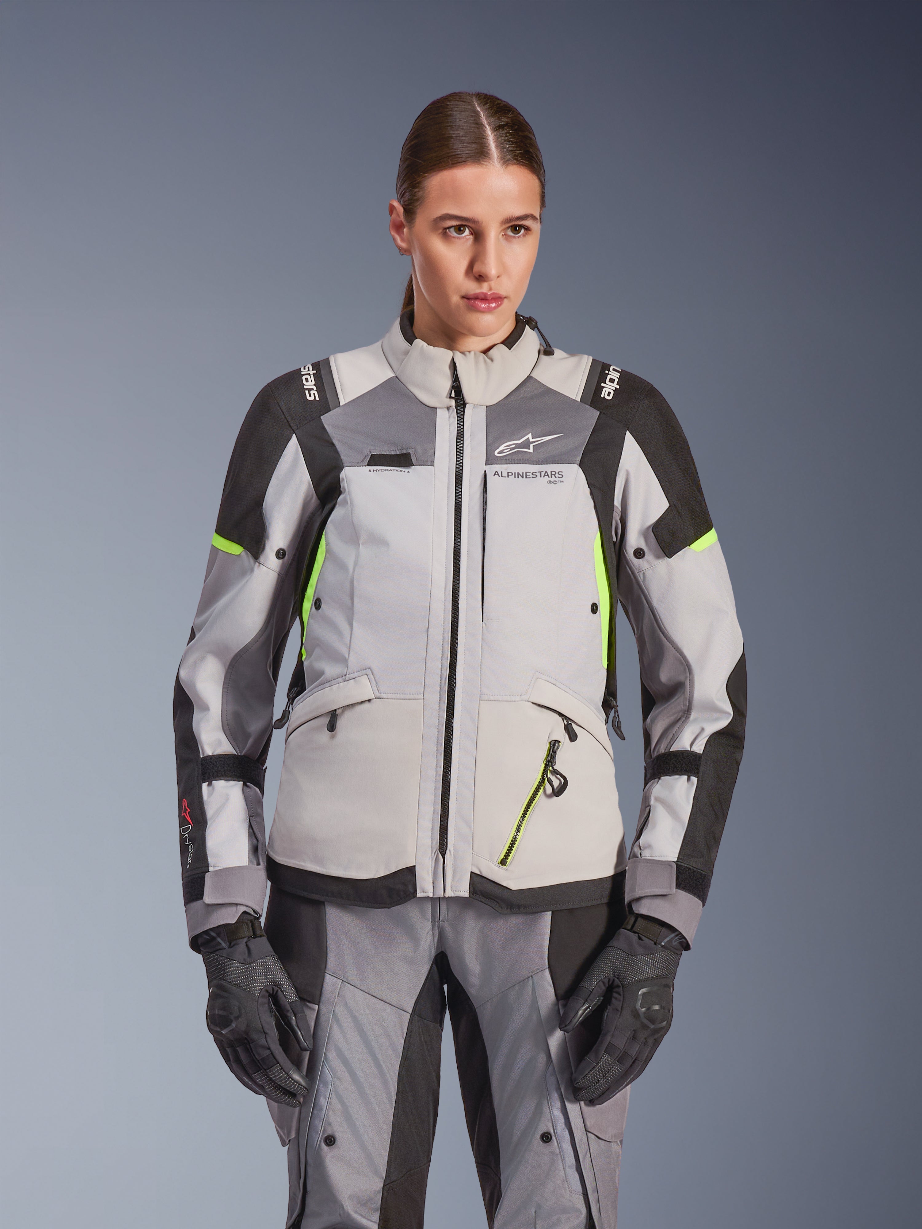 Alpinestars Stella Andes V4 Drystar® Damenjacke, Eisgrau und Dunkelgrau mit neongelben Akzenten, Adventure-Touring-Motorradjacke, getragen mit passender Hose und Handschuhen, schützende Textilkonstruktion mit Alpinestars-Branding.