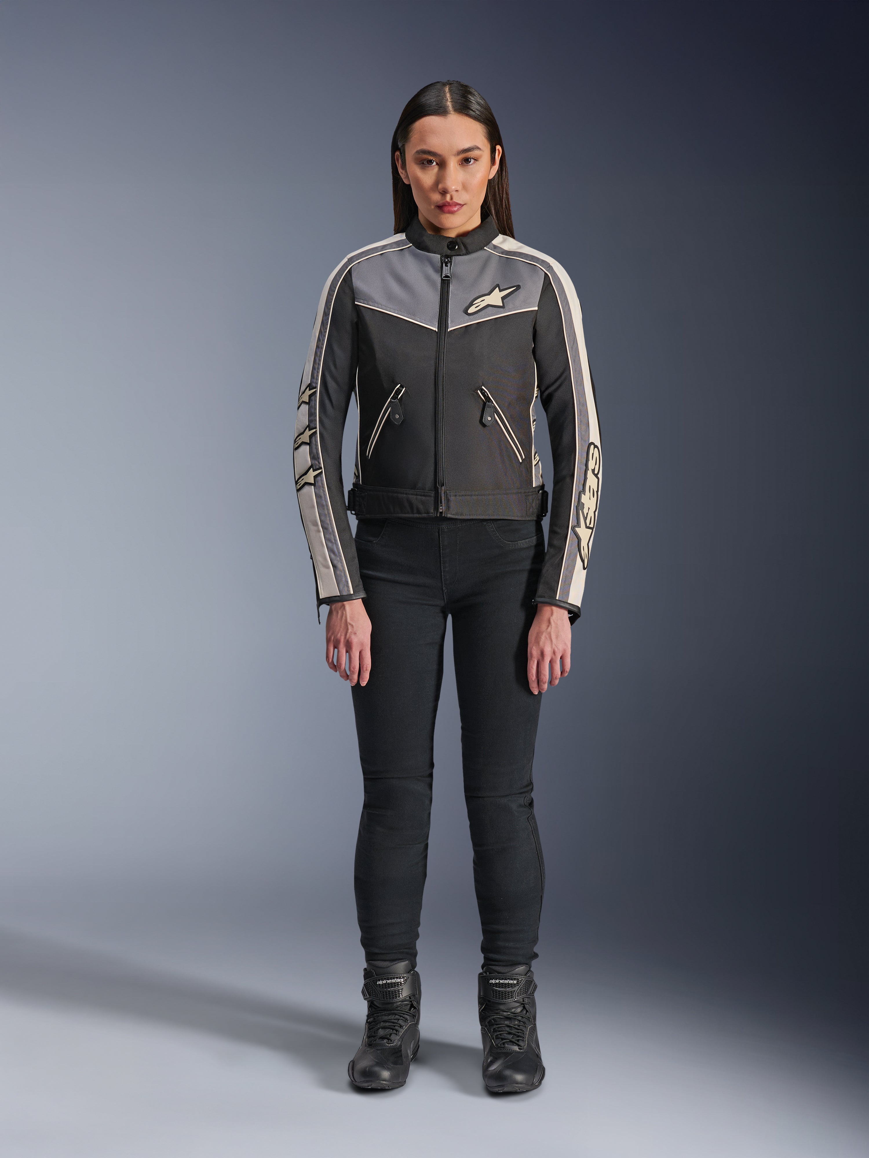 Frau in Alpinestars Stella T-Dyno WR Jacke, Schwarz, Dunkelgrau und Ecru, Vorderansicht, Textil-Motorradjacke mit vintage-inspiriertem Design, Branding an Brust und Ärmeln, getragen mit schwarzer Hose und Stiefeln