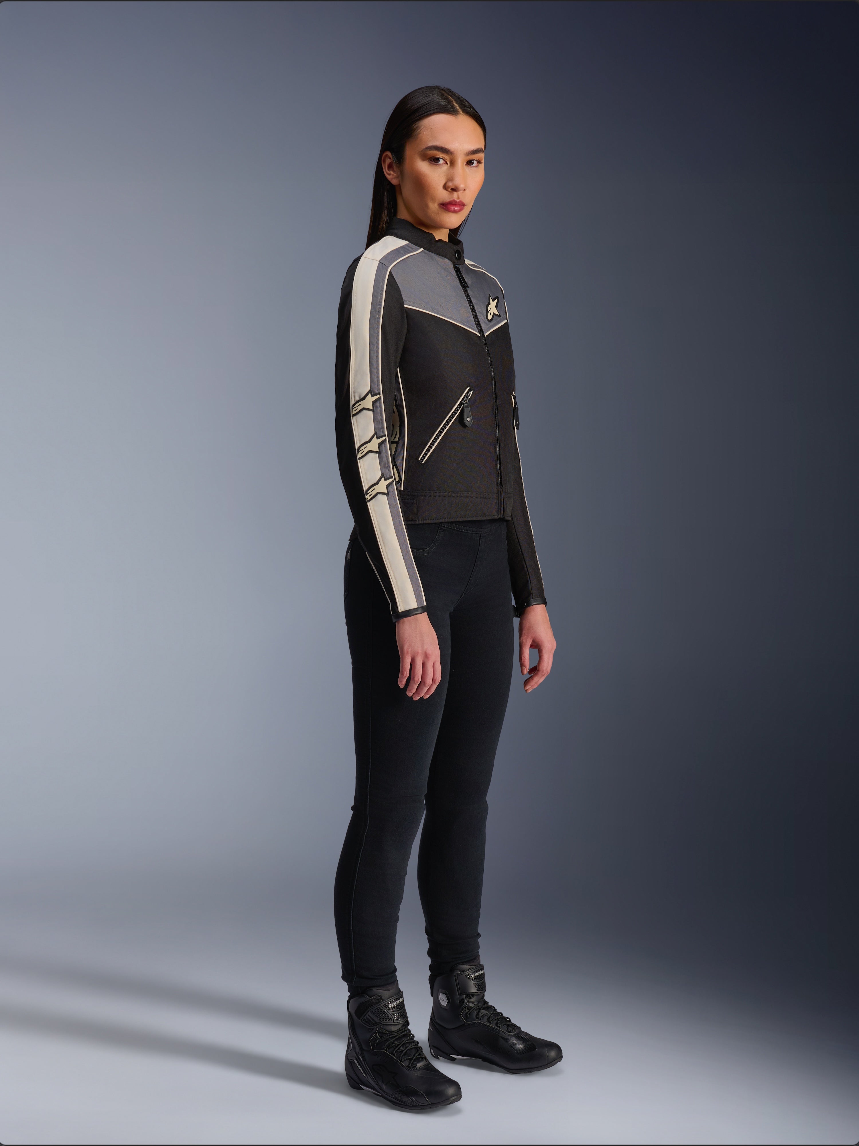 Alpinestars Stella T-Dyno WR Jacke, Textil-Motorradjacke für Damen, Schwarz, Dunkelgrau und Ecru, Vintage-inspiriertes Design mit cremefarbenen Ärmelstreifen und Stern-Logos, getragen mit schwarzer Hose und Motorradstiefeln, Heritage-inspirierte Urban-Riding-Ausrüstung