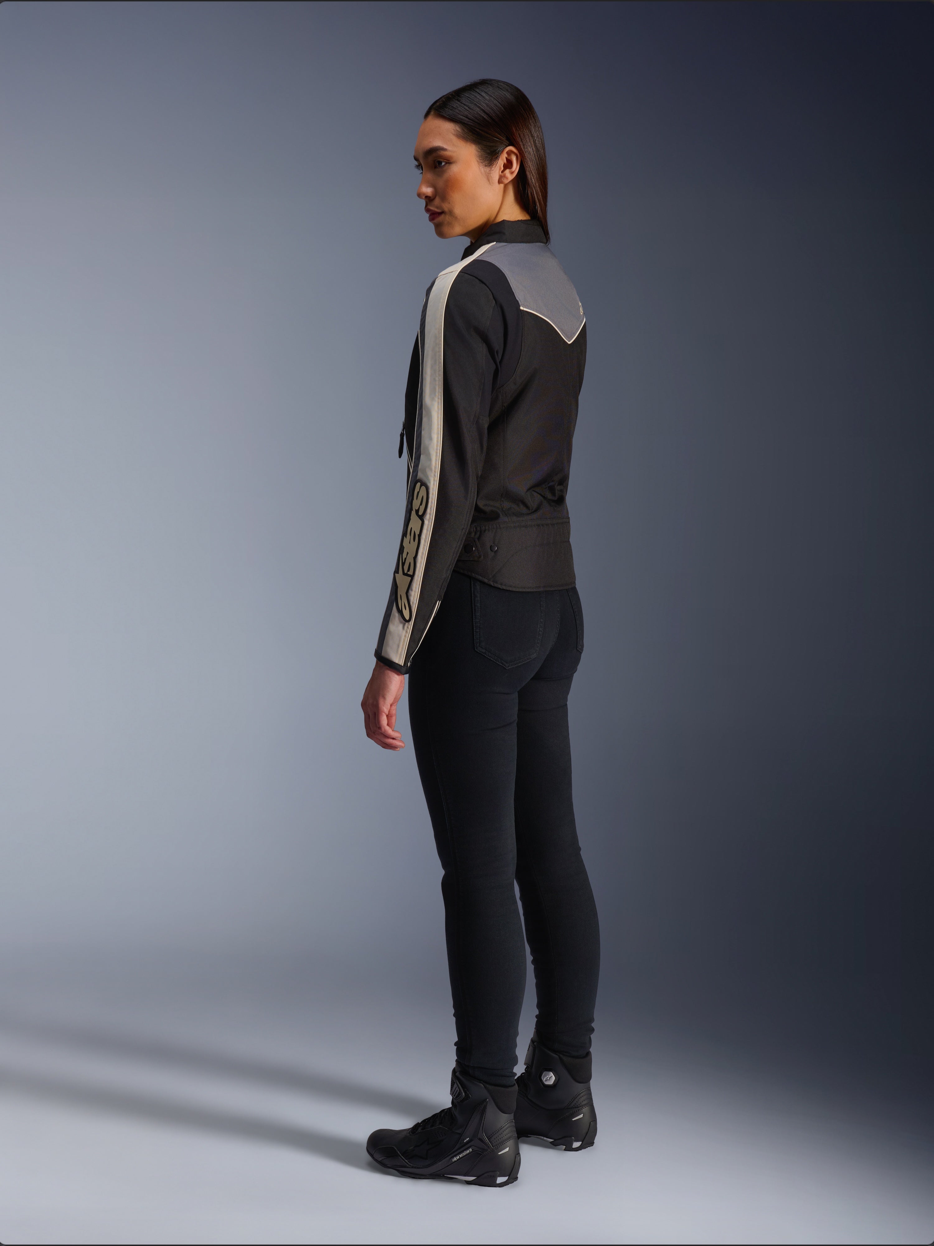 Weibliches Model in der Seiten- und Rückansicht, trägt die Alpinestars Stella T-Dyno WR Jacke in Schwarz Dunkelgrau Ecru, mit vintage-inspiriertem Design, ecrufarbenen Ärmelstreifen und grauer Rückenpartie, kombiniert mit schwarzer Hose und schwarzen Motorradschuhen.