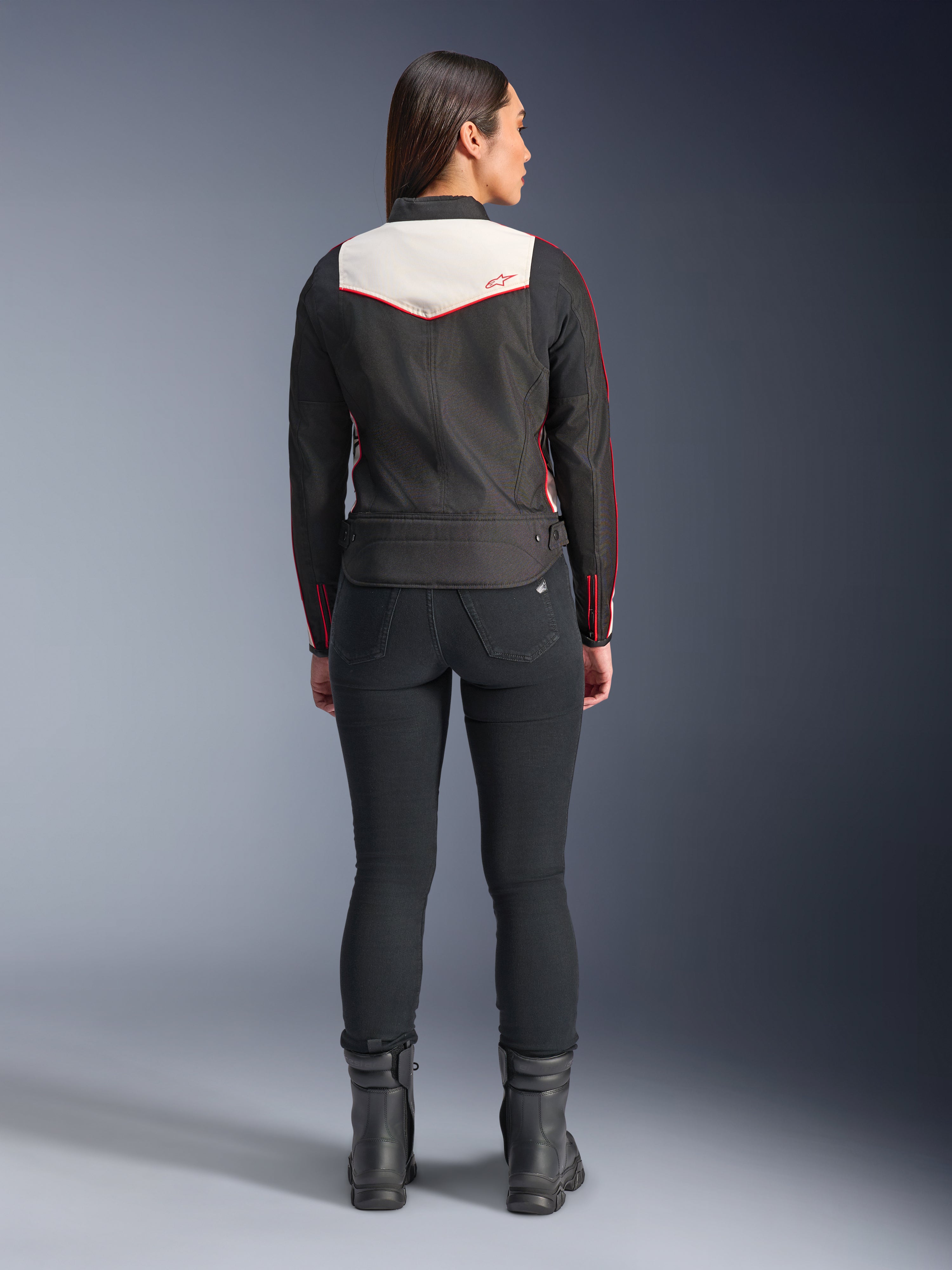 Rückansicht eines weiblichen Models in der Alpinestars Stella T-Dyno WR Jacke in Black Ecru Bright Red, mit einem markanten cremefarbenen Rückenpanel und roten Logo-Akzenten, kombiniert mit einer schwarzen Hose und Stiefeln, eine vintage-inspirierte Textil-Motorradjacke.
